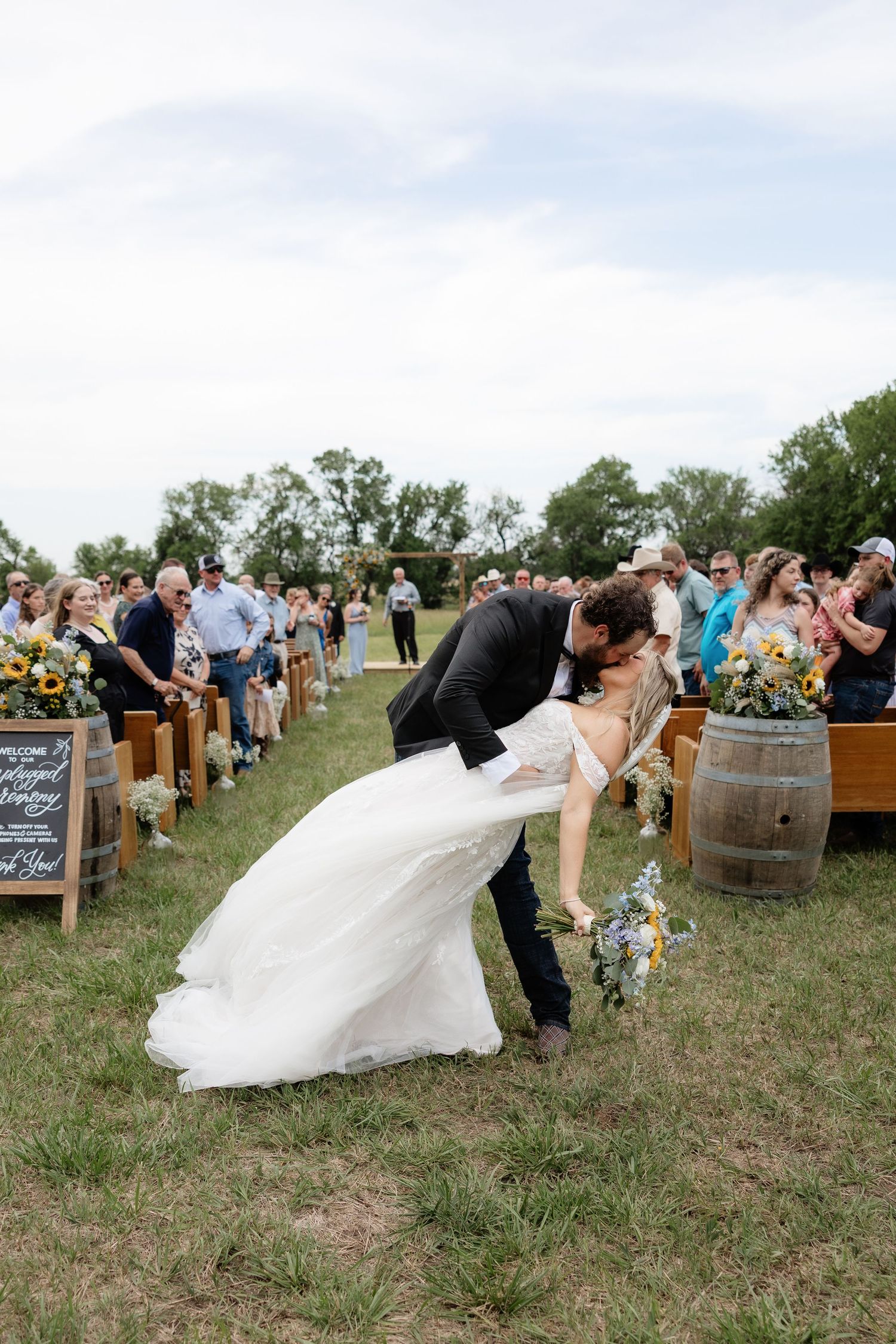 Groom dips bride for romantic kiss in aisle at outdoor rustic farm wedding celebration.