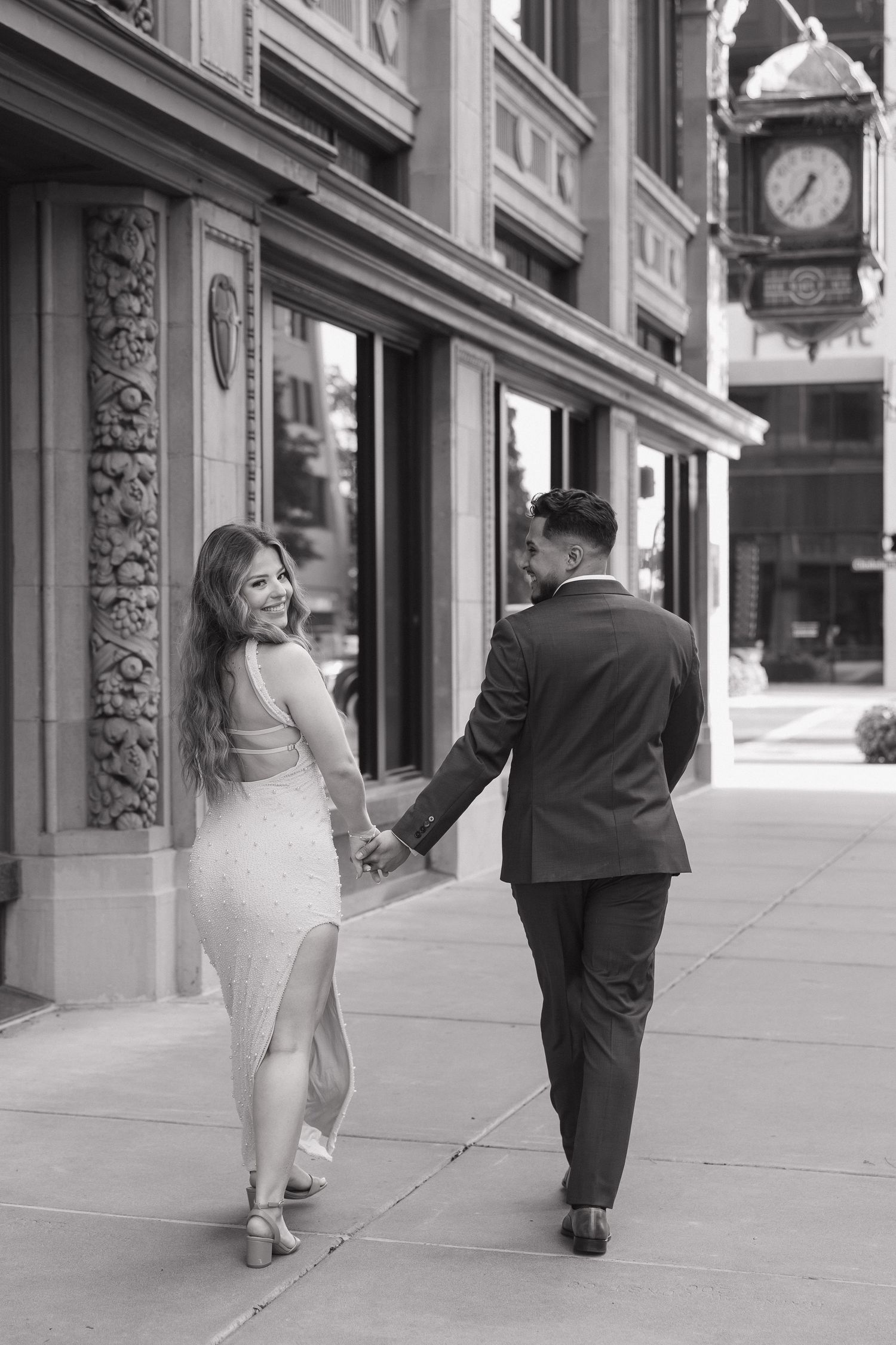 Black and white photo of couple holding hands walking past ornate building with vintage street clock.