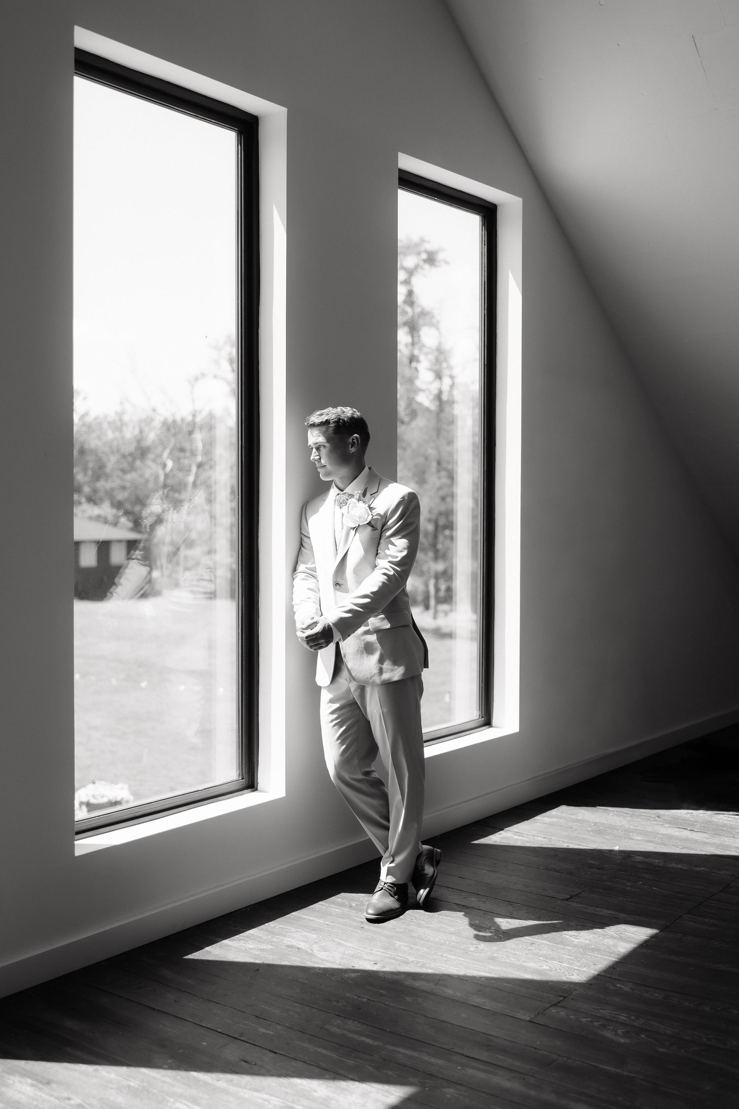 Black and white portrait of a groom in a light suit standing by large windows in dramatic sunlight.