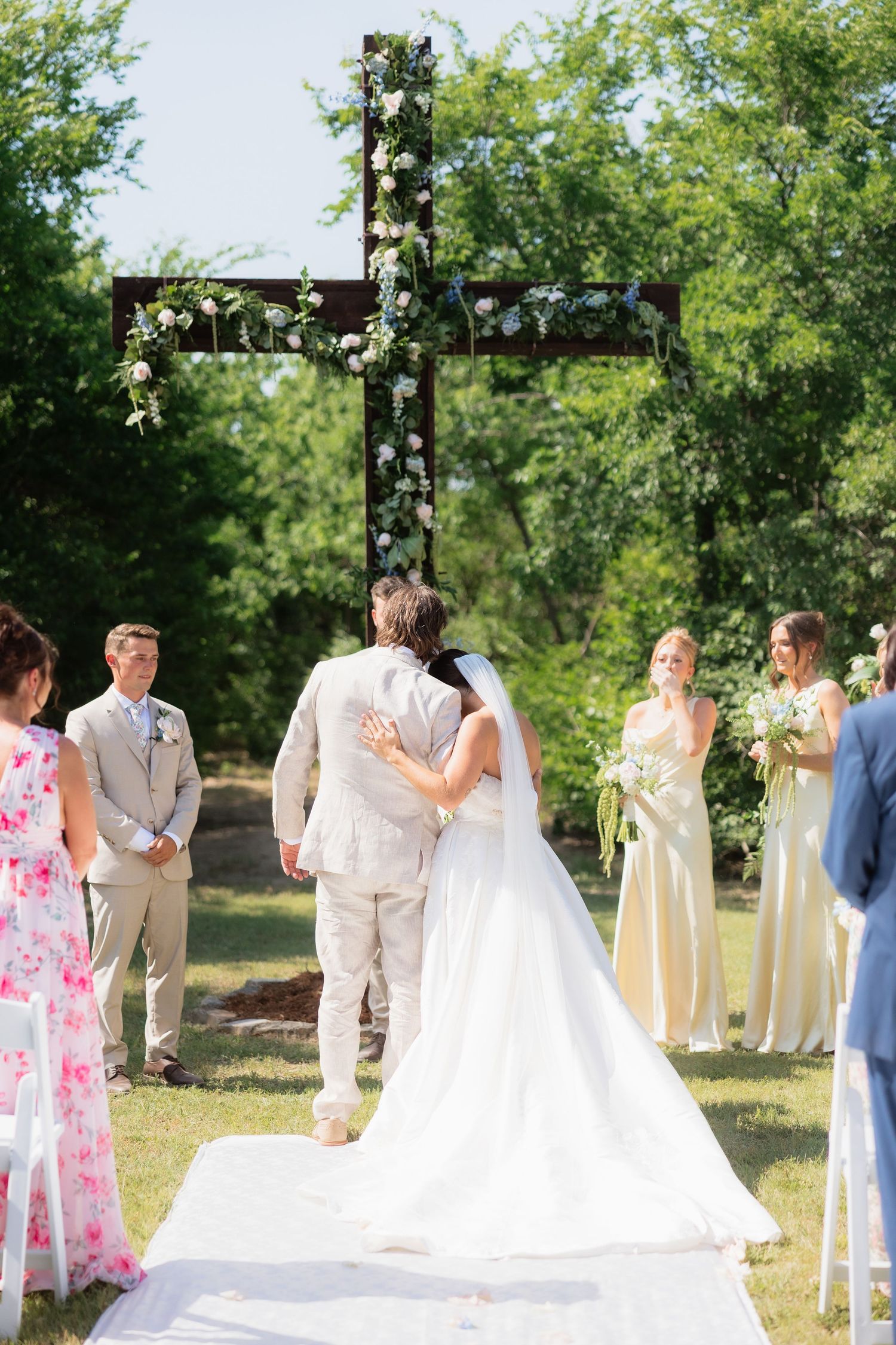 Bride and groom embrace at outdoor wedding altar adorned with floral cross surrounded by bridesmaids.
