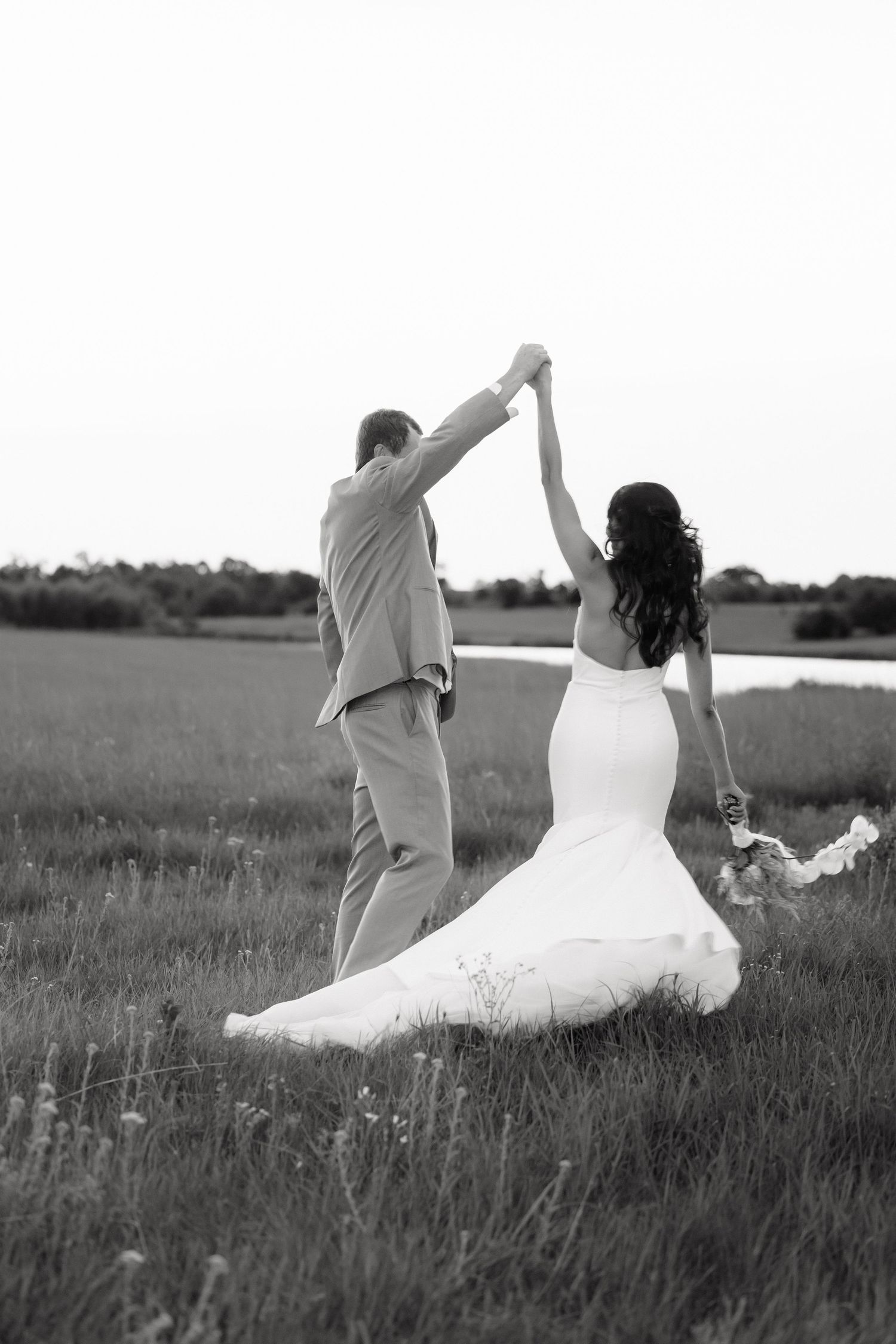 Black and white photo of bride and groom dancing in open field, hands raised joyfully.