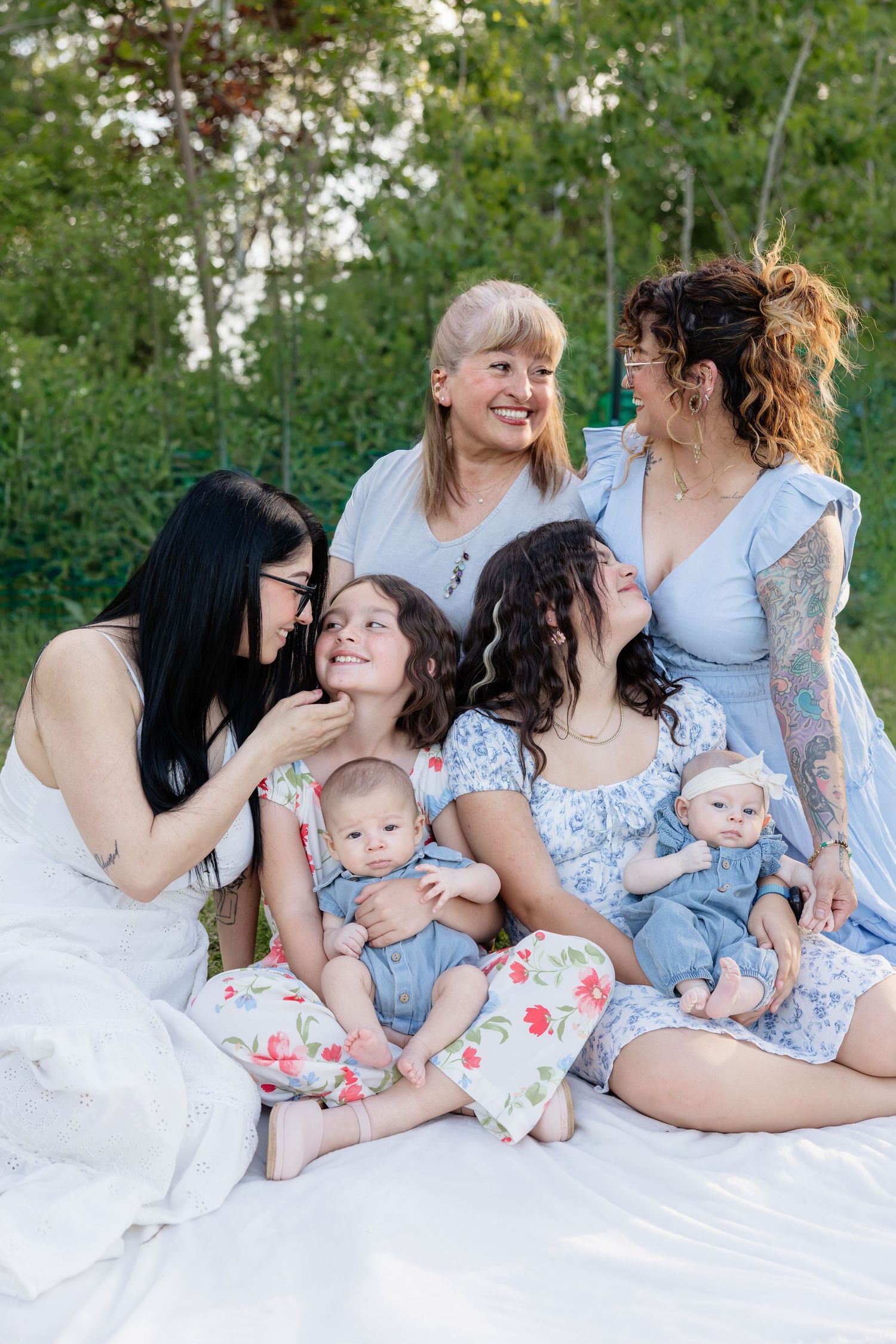 A multigenerational family group sits on a white blanket outdoors, smiling and holding babies in a garden.