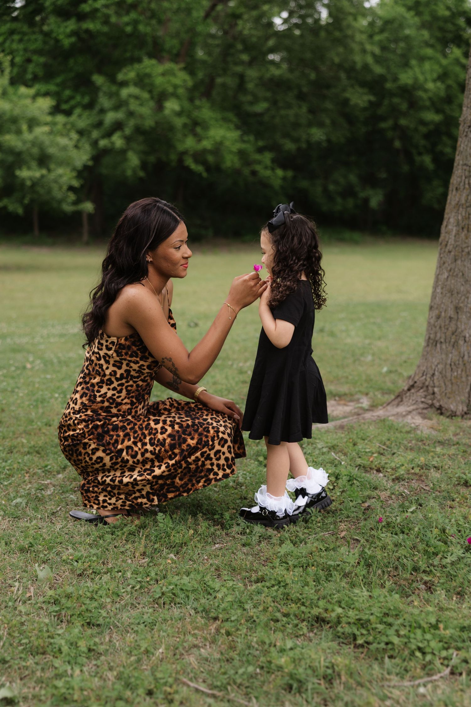 A woman in leopard print dress kneels to offer a flower to a curly-haired toddler in a park.