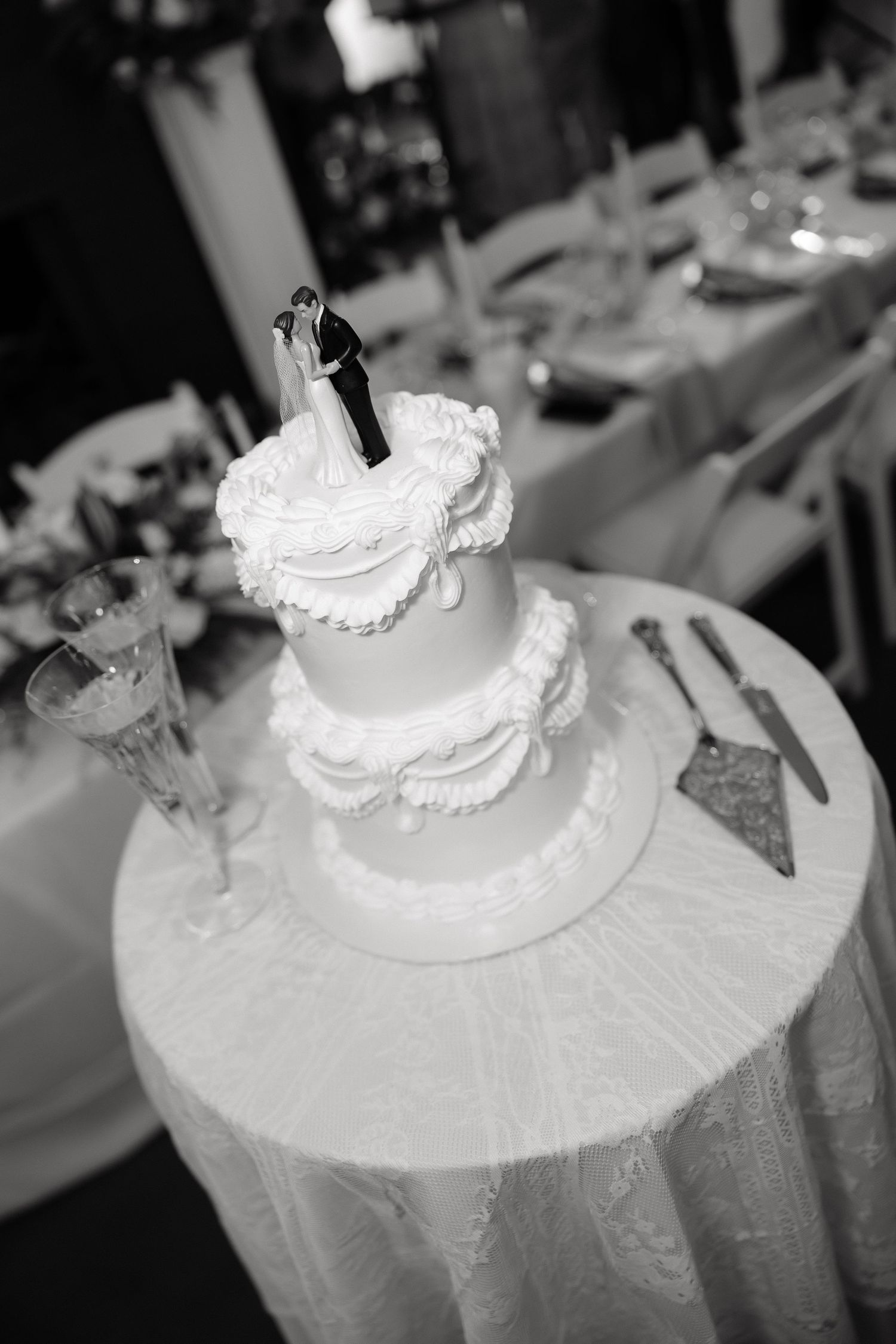 Black and white photo of two-tier wedding cake with bride and groom topper displayed on decorated reception table.