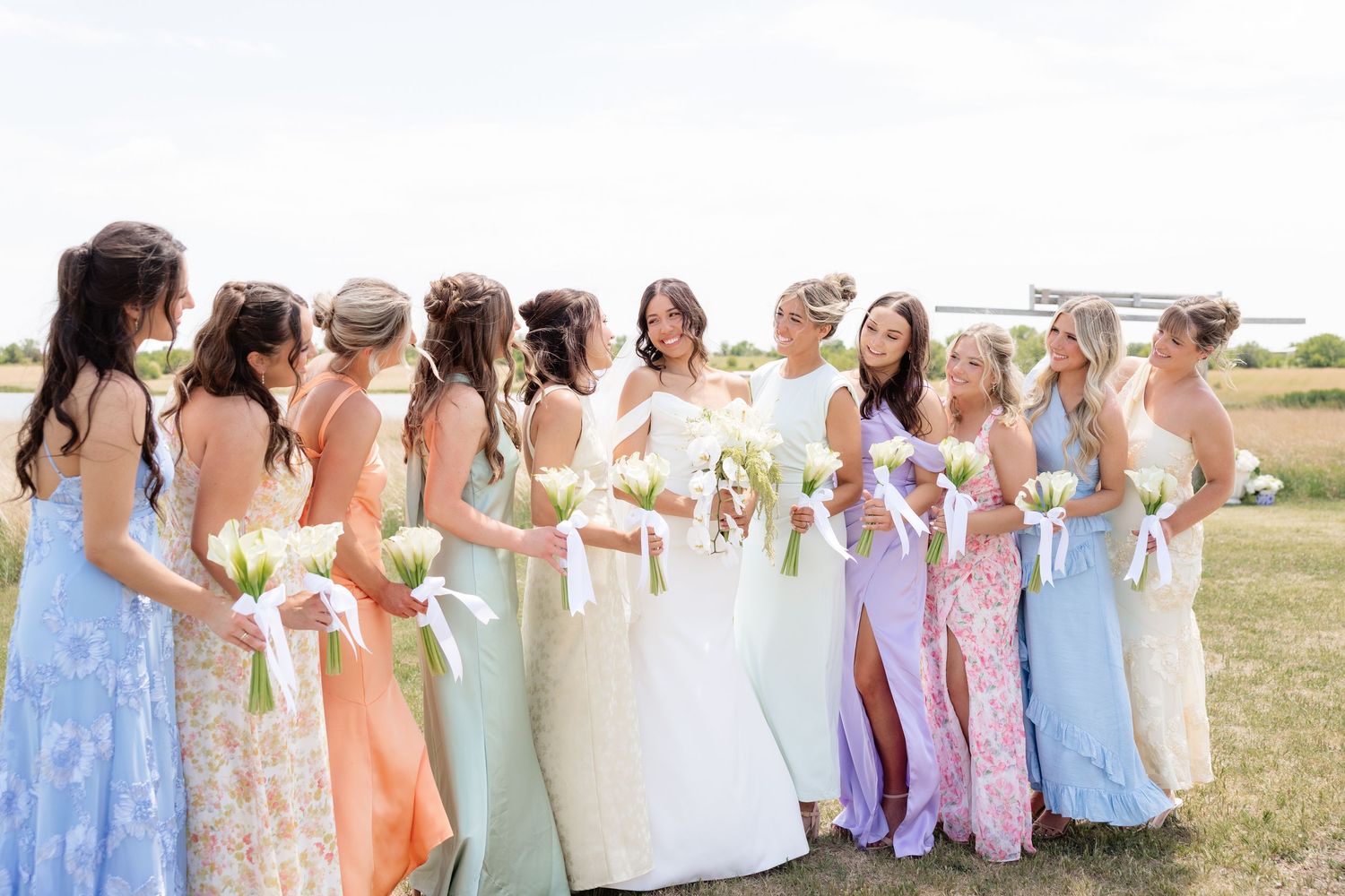 Bride smiles with large bridal party in colorful mismatched gowns at outdoor wedding.