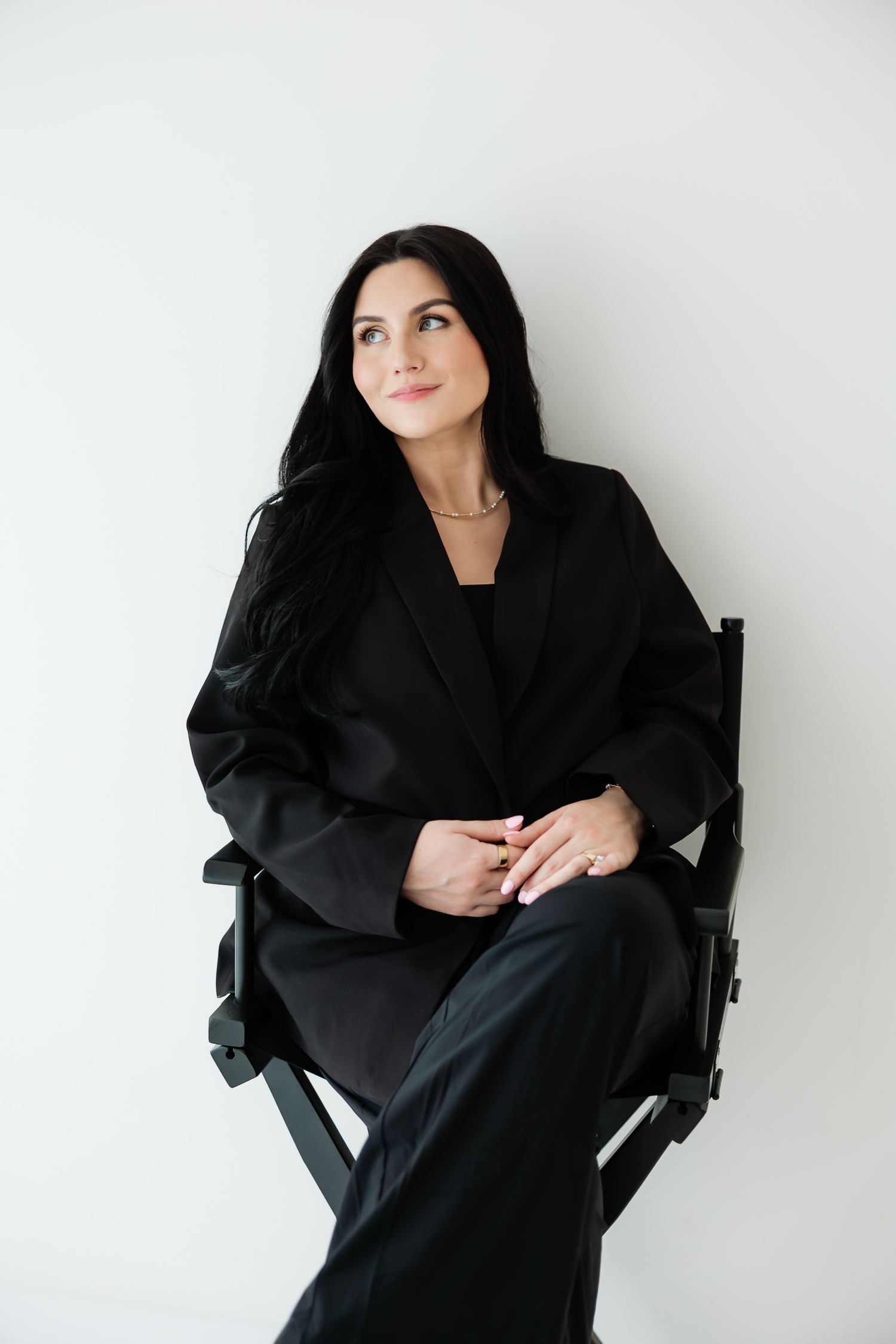 Brunette woman in black blazer sitting on director's chair against white wall.
