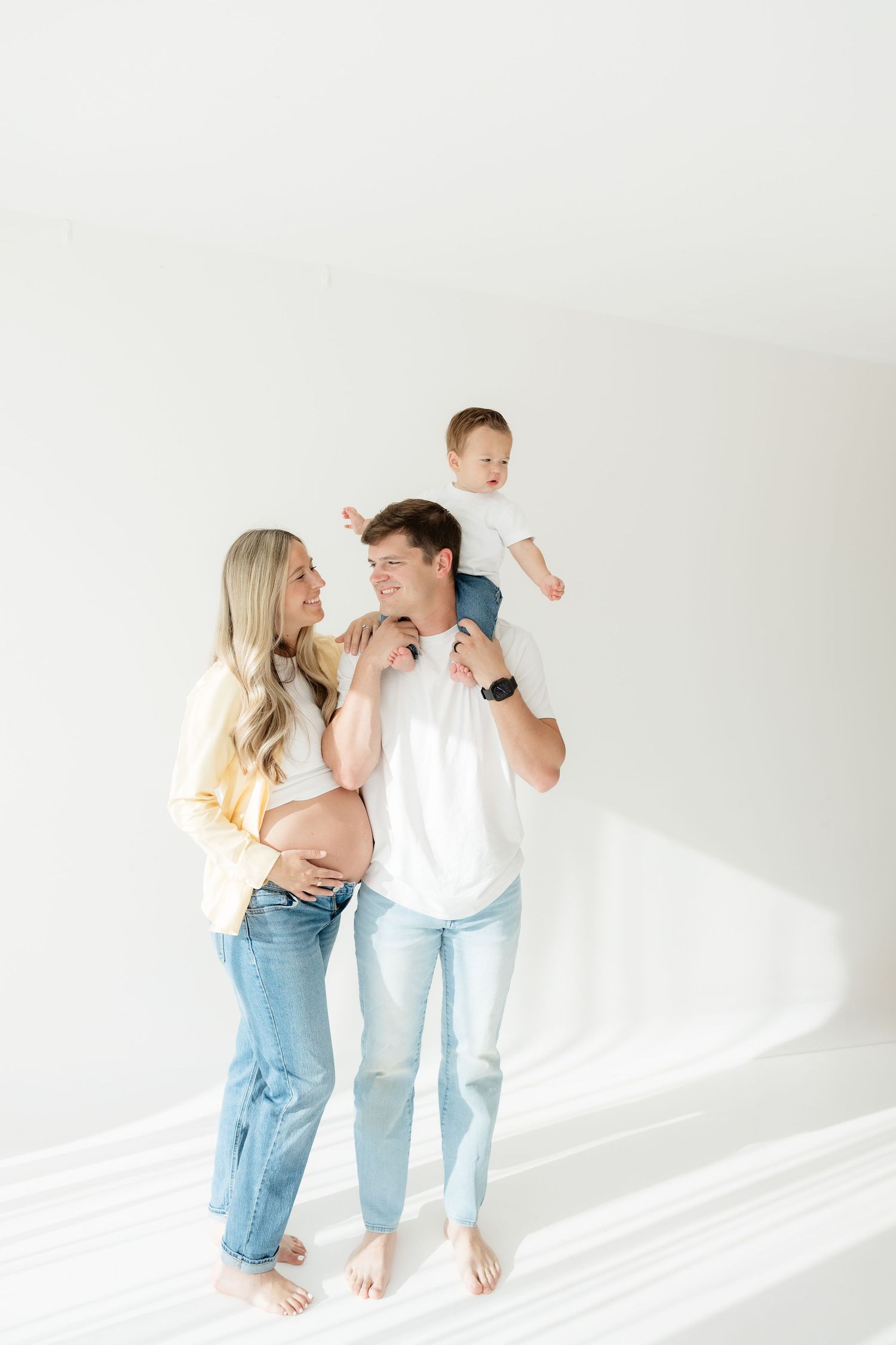 A happy pregnant woman and man in jeans hold a baby on his shoulders in a bright white studio setting.
