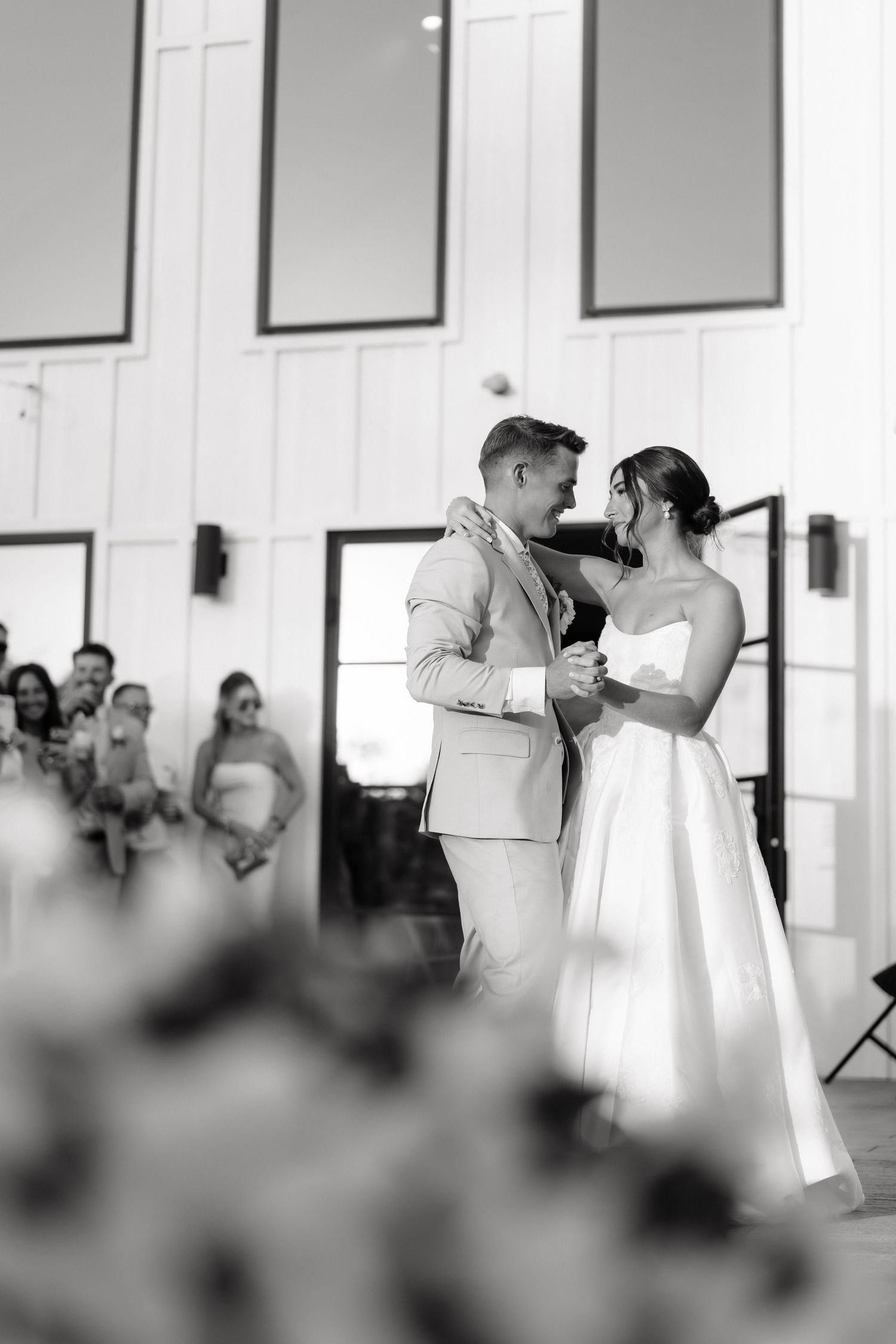 Black and white photo of bride and groom sharing their first dance inside a modern white barn venue.