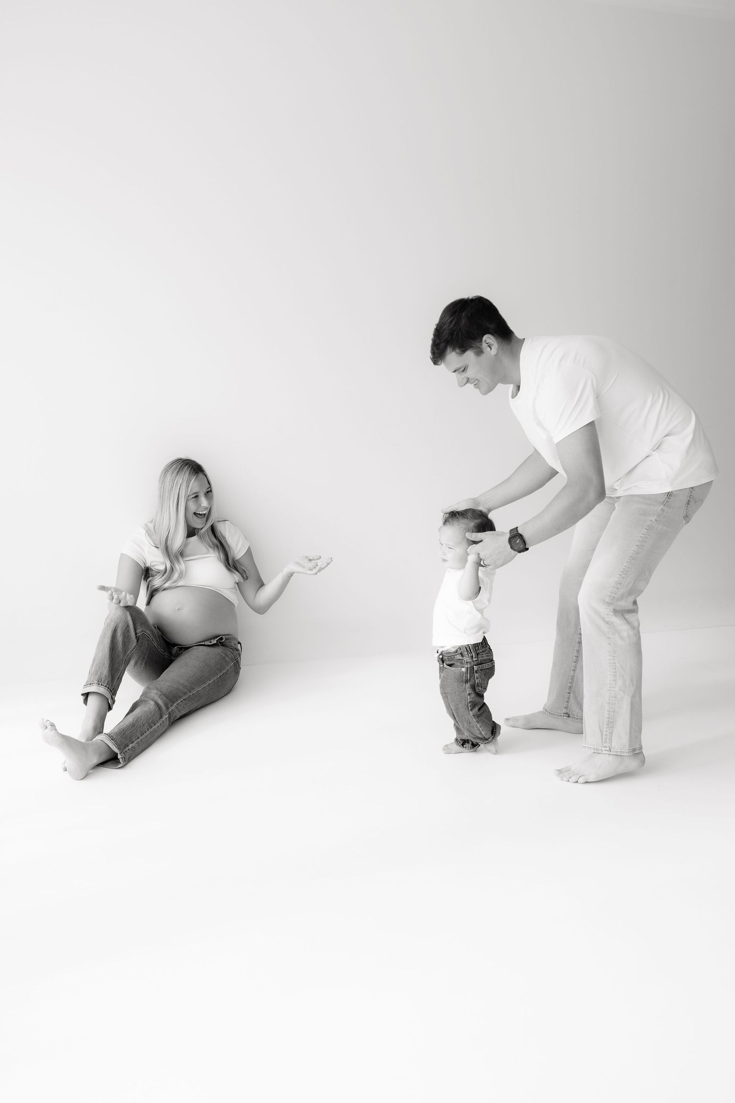 Black and white photo of pregnant woman sitting on floor as husband helps toddler walk toward her.