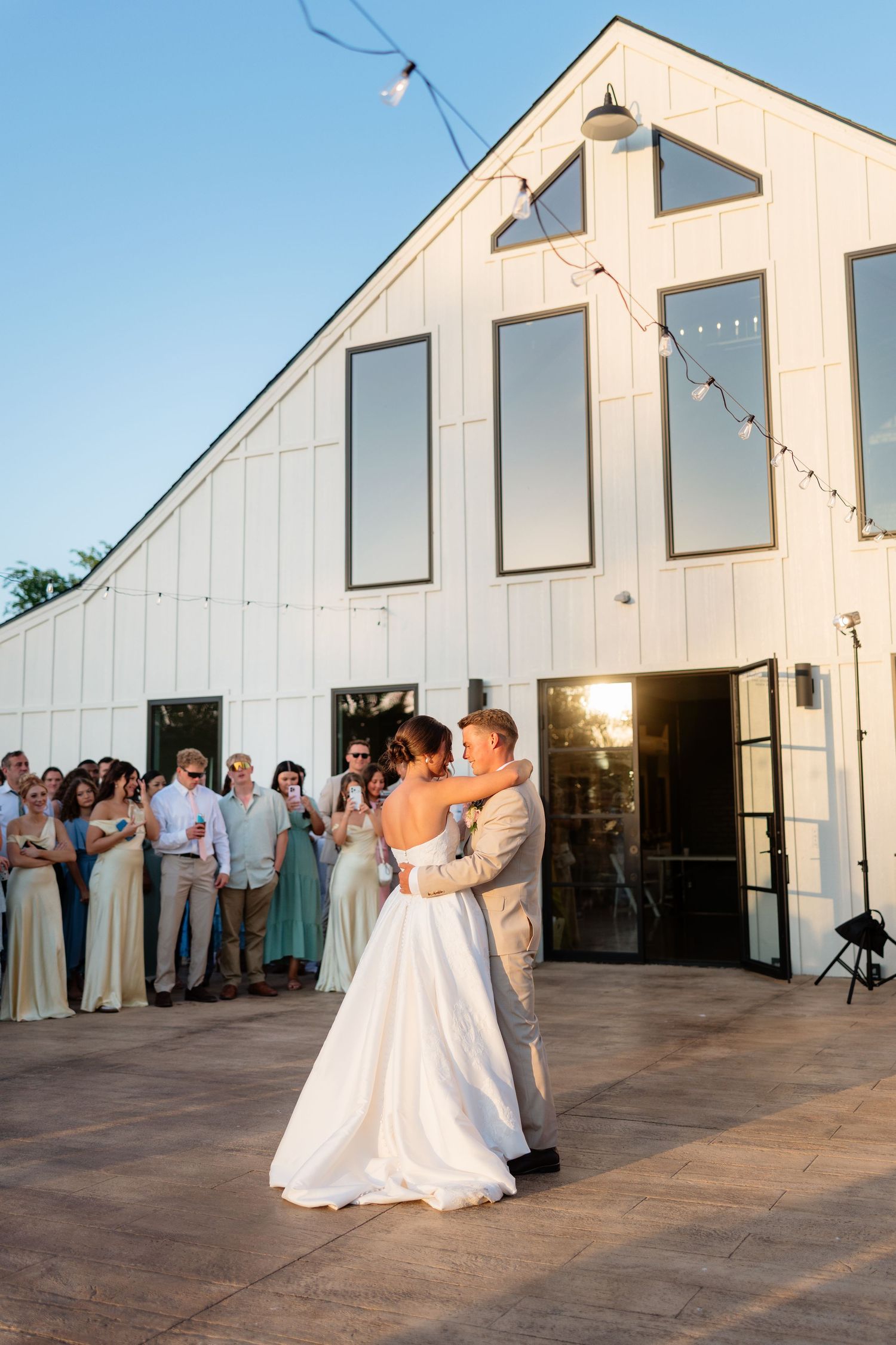 Bride and groom dancing outdoors at sunset in front of a white barn venue surrounded by wedding guests.