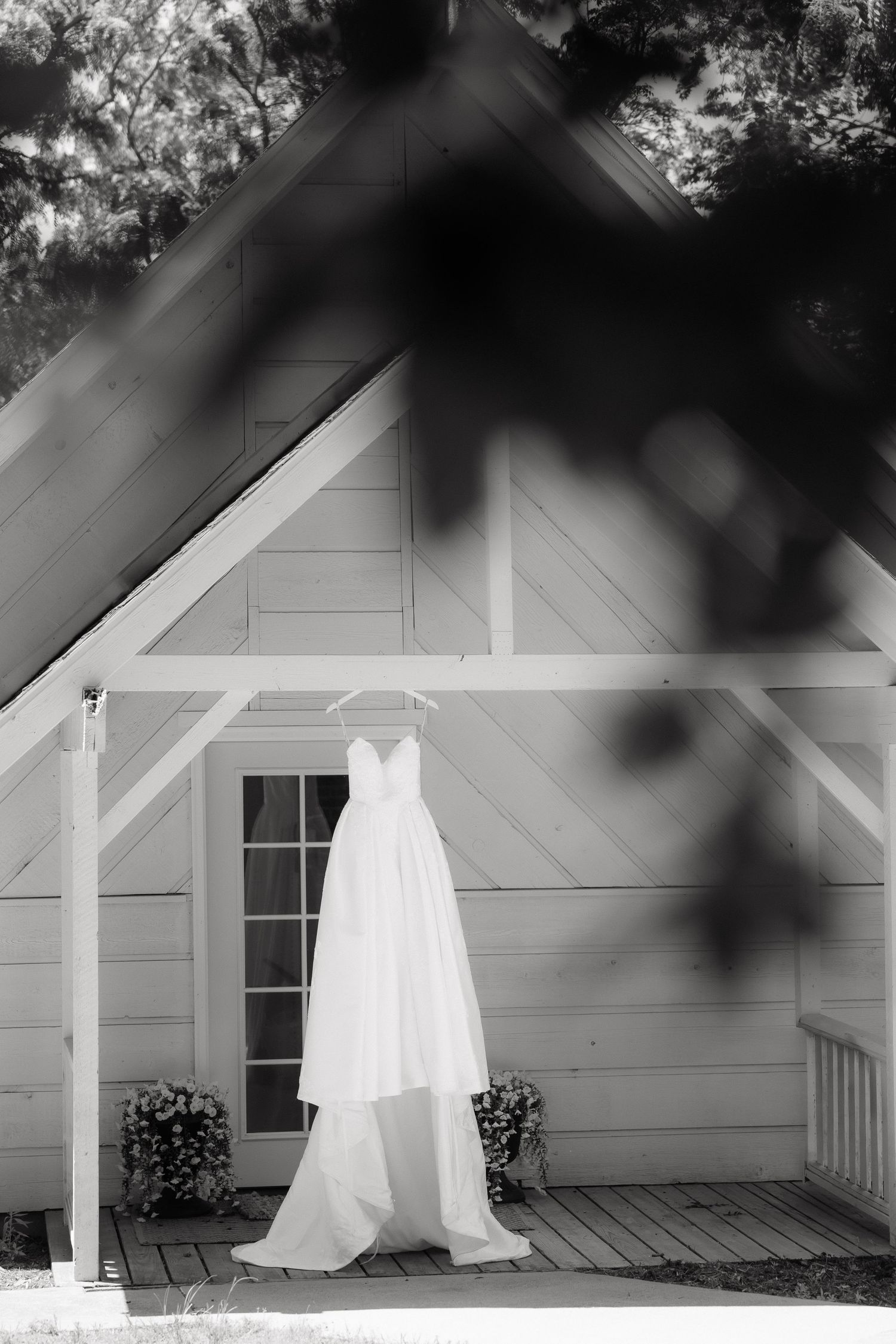 Black and white photo of strapless wedding gown hanging on wooden chapel exterior.