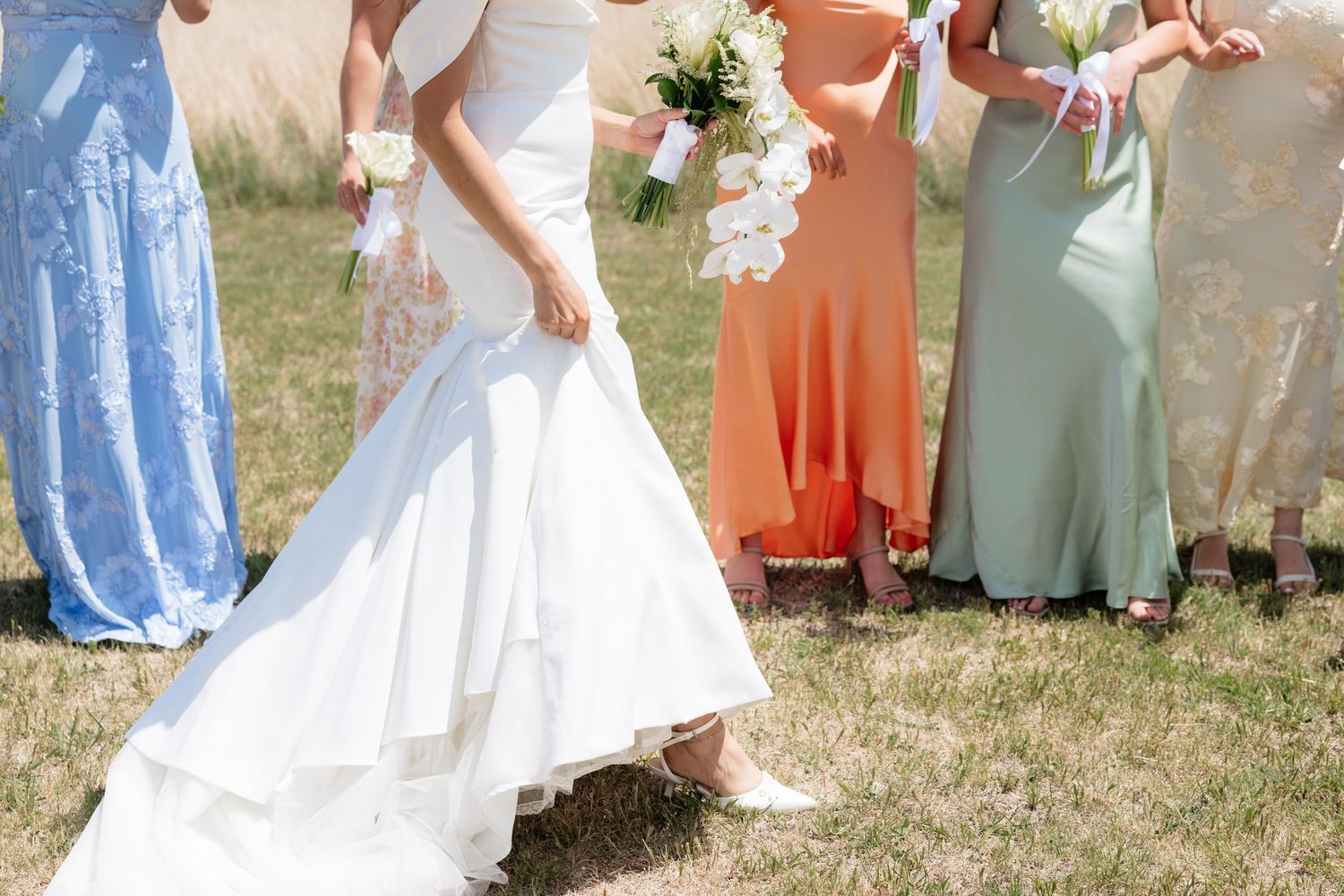 Bride in mermaid gown walks with colorful bridesmaids holding white bouquets outdoors.