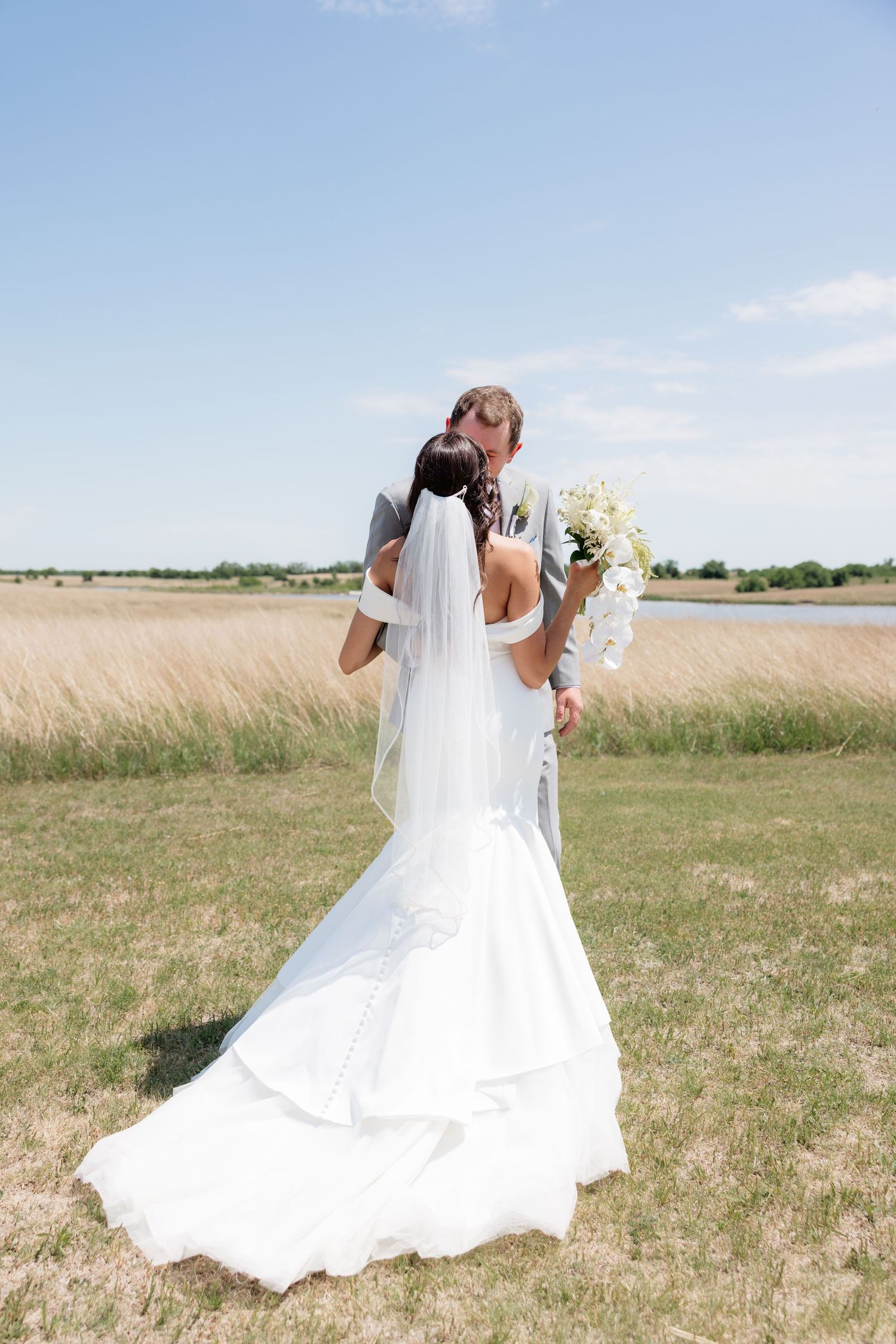 Bride and groom share a kiss in a sunny field, bride holding white orchid bouquet with veil trailing.