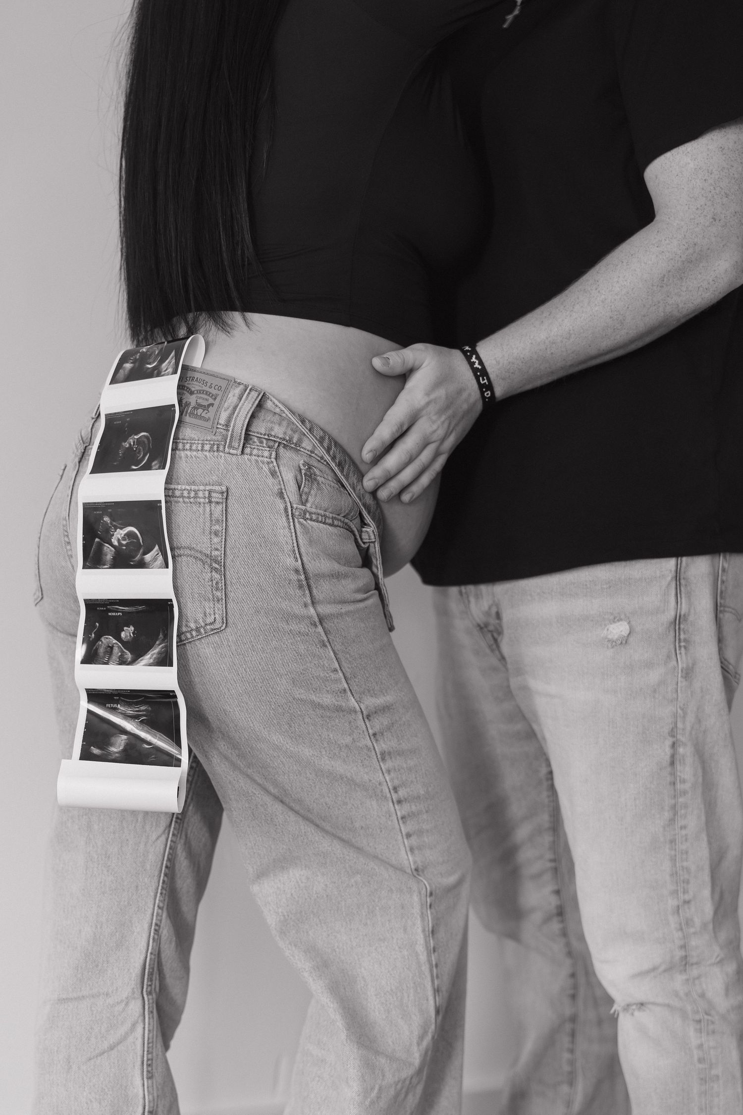 Black and white maternity photo of a couple with ultrasound images tucked into the mothers unbuttoned jeans.