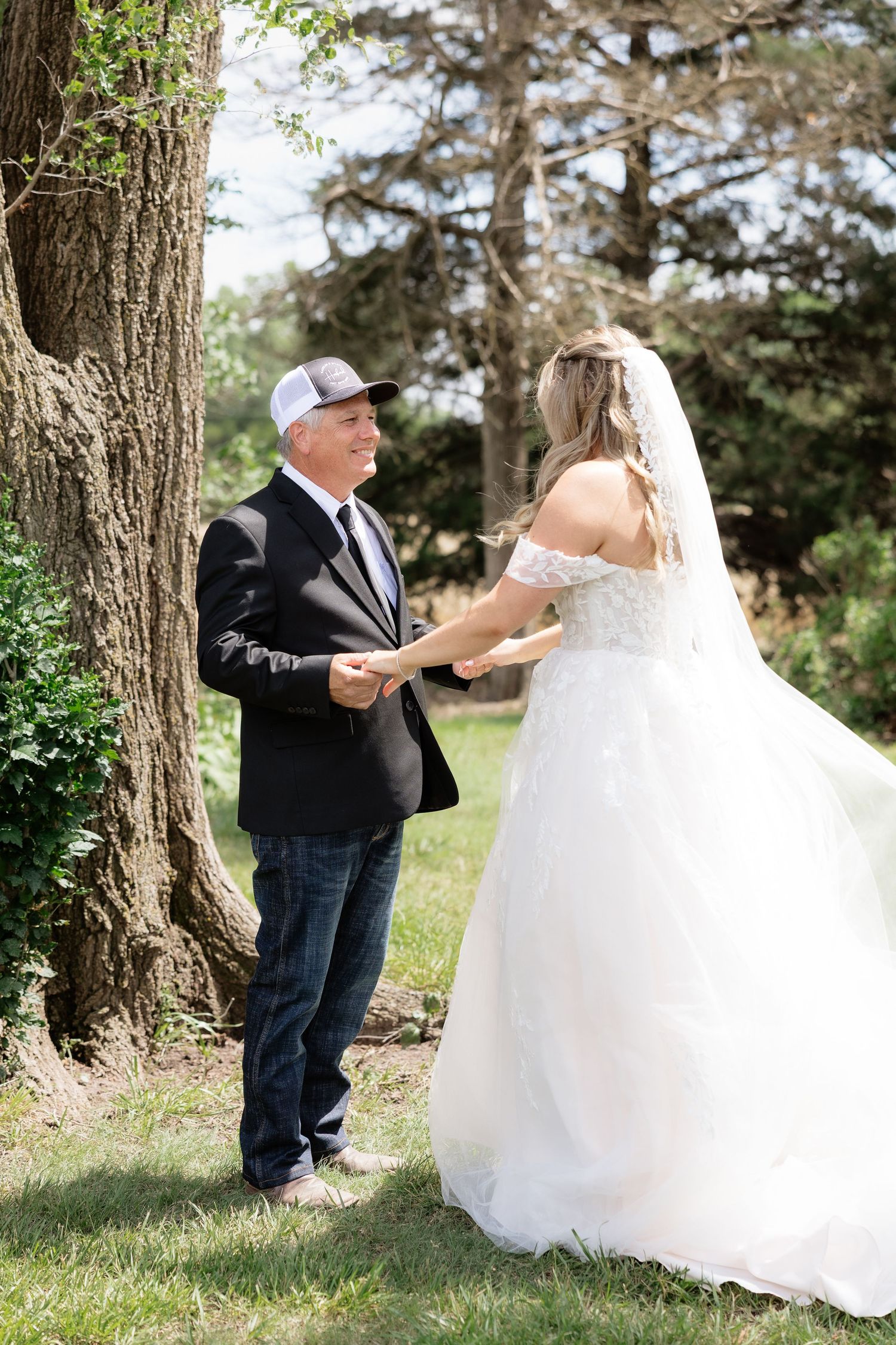 Father of the bride holds daughter's hands during emotional first look reveal near a large tree outdoors.