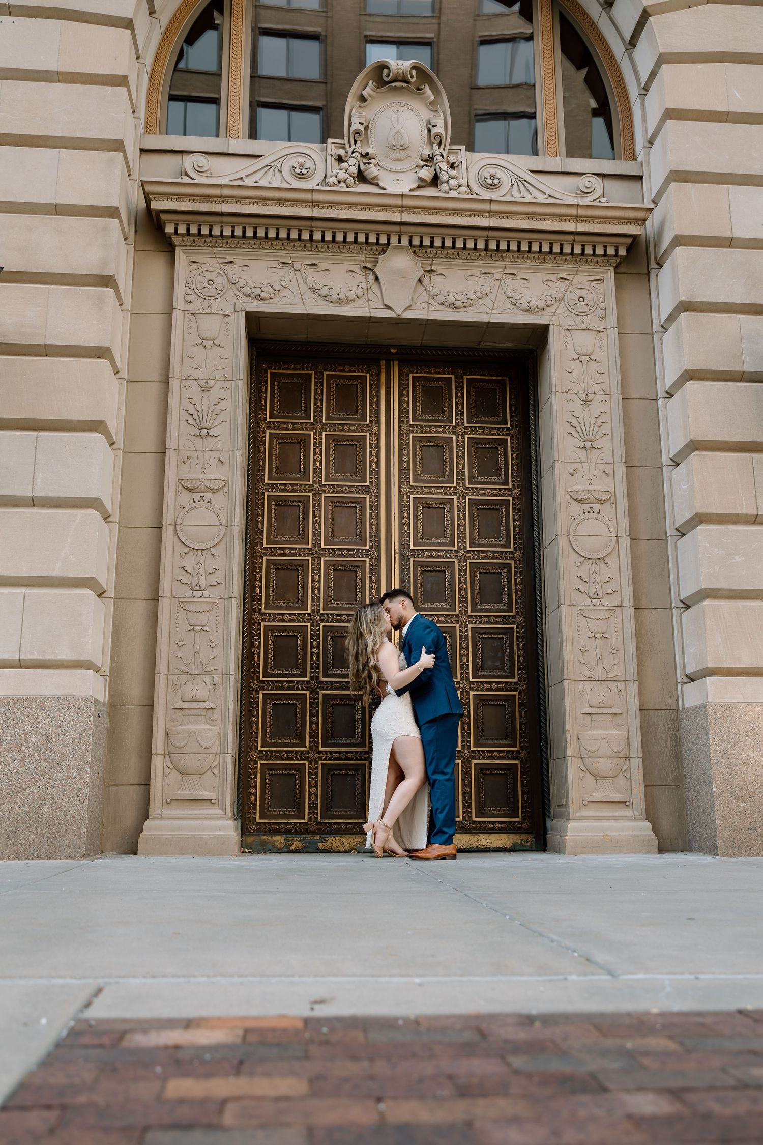 Engaged couple kissing before grand ornate bronze doors of a classical stone building.