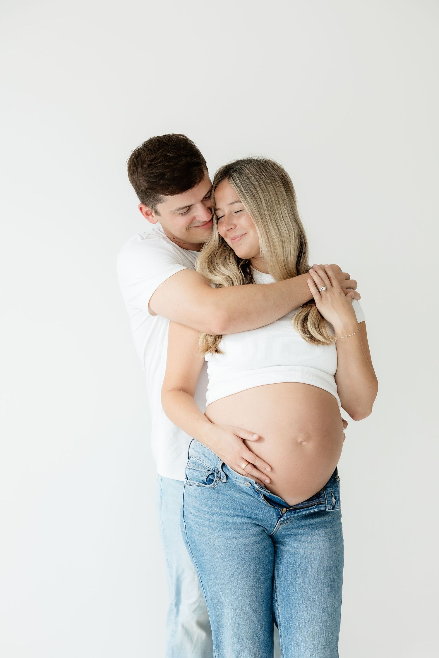 Man embraces pregnant woman from behind as she holds baby bump against white studio background.
