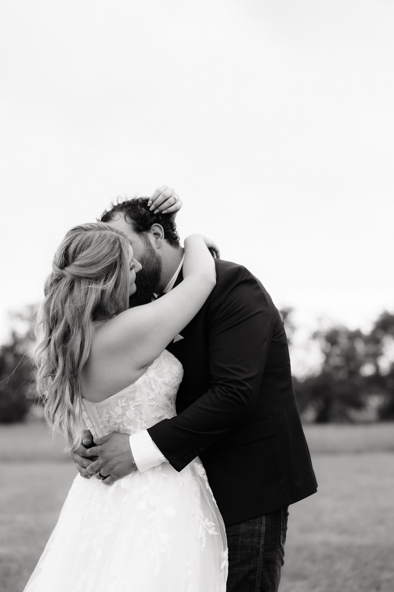 Black and white photo of bride and groom embracing closely in open field after wedding ceremony.