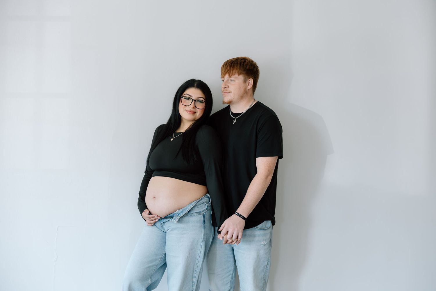 Expecting couple in matching black tops and jeans posing against a white wall during maternity photoshoot.