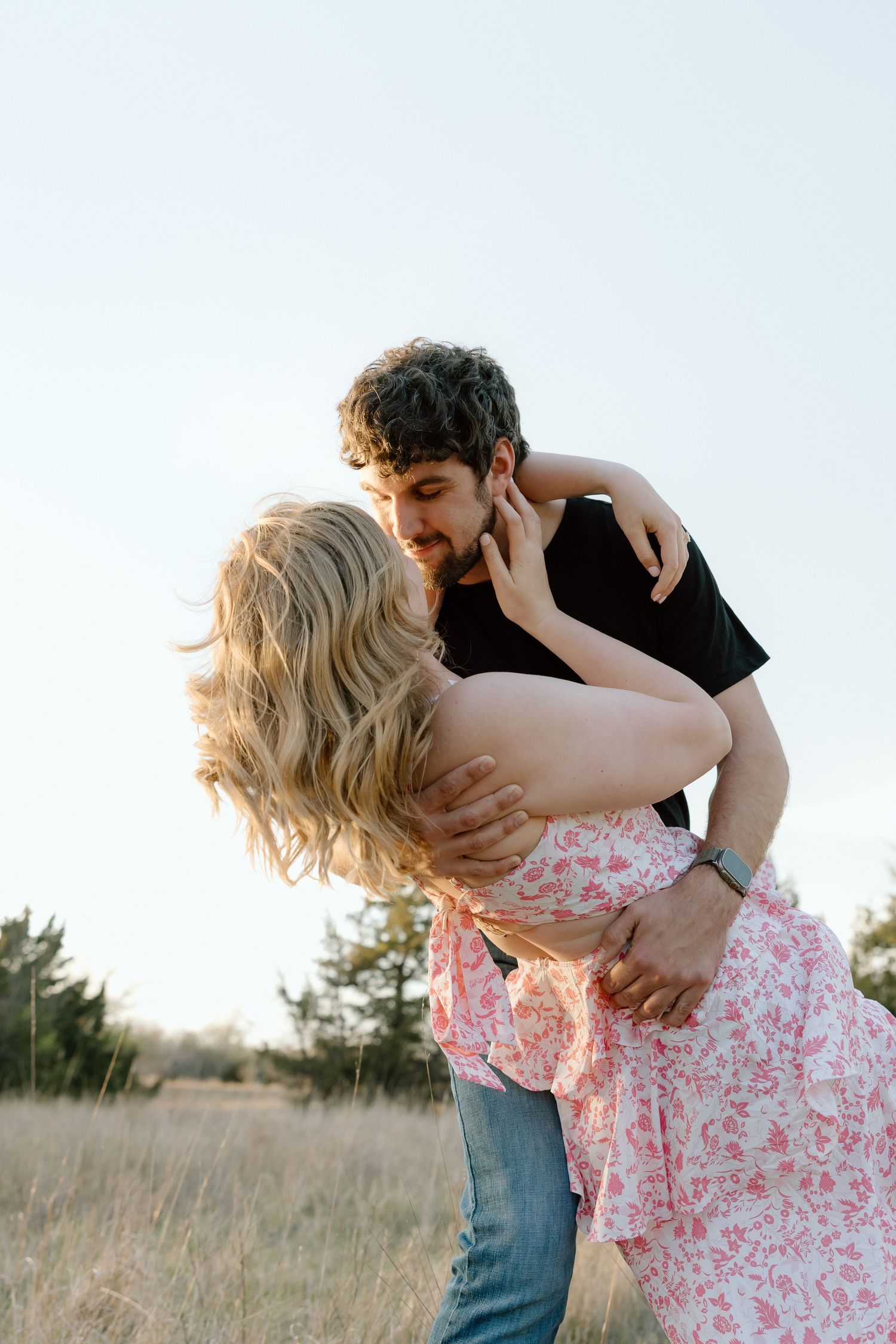 Couple sharing a romantic dip kiss in an open field during golden hour, woman in floral pink dress.