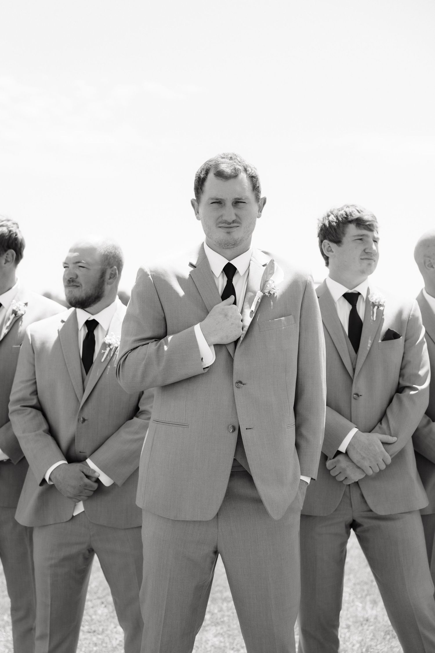 Black and white portrait of groom adjusting boutonniere surrounded by groomsmen in gray suits outdoors.