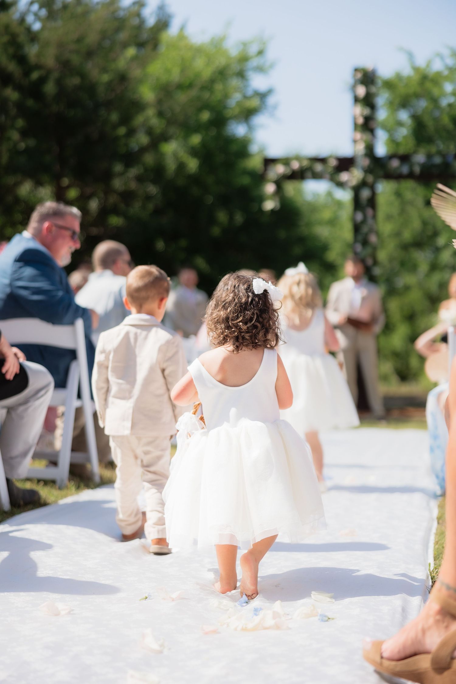 Flower girl in white dress and ring bearer walk down aisle at outdoor wedding ceremony with floral cross.