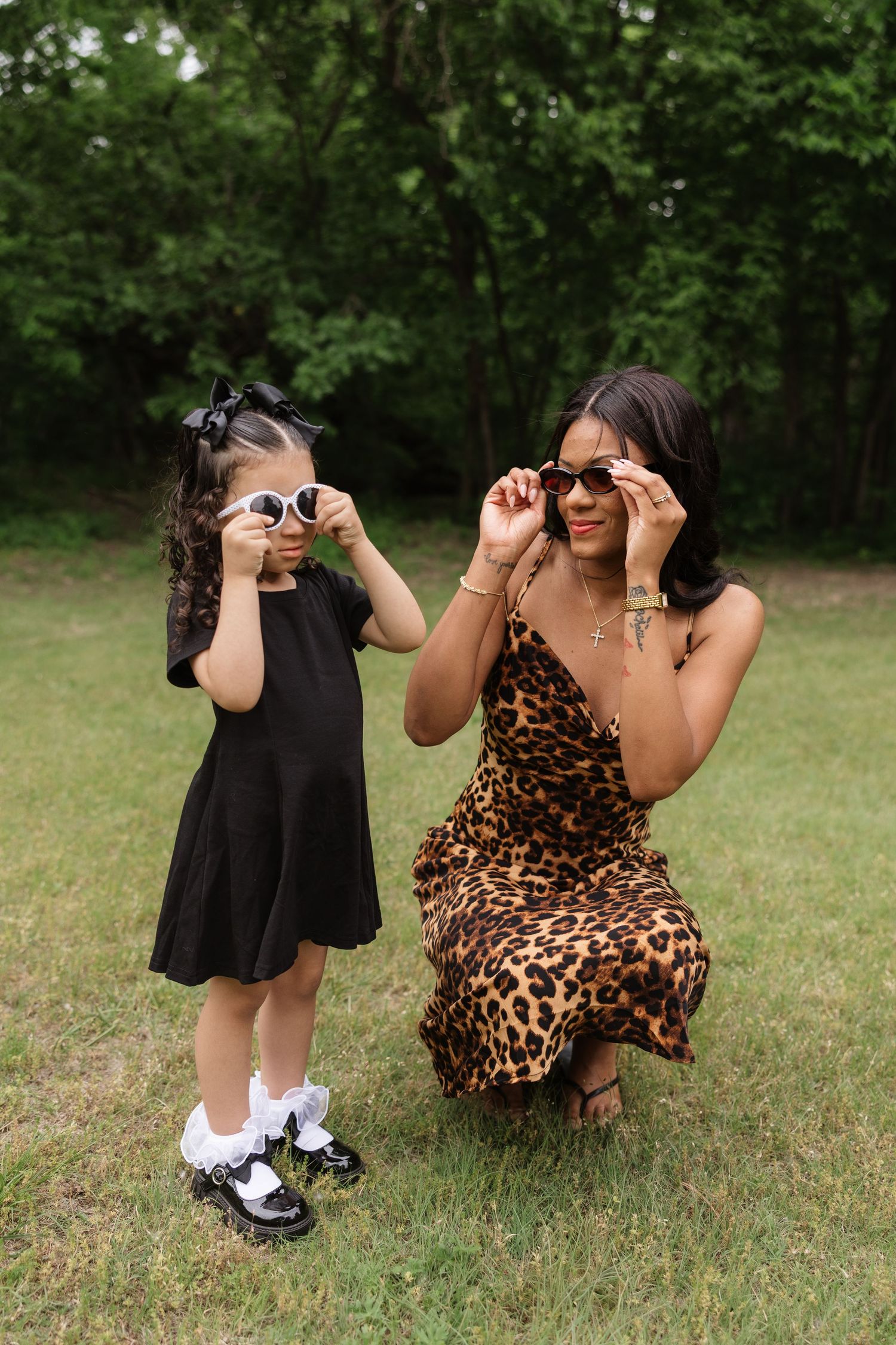 Mother and daughter adjusting matching sunglasses together on a green lawn.