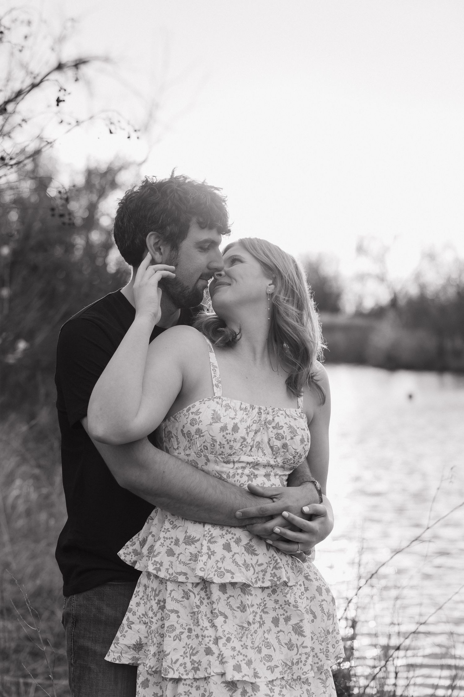 Black and white photo of a romantic couple embracing and almost kissing near a serene lake at sunset.