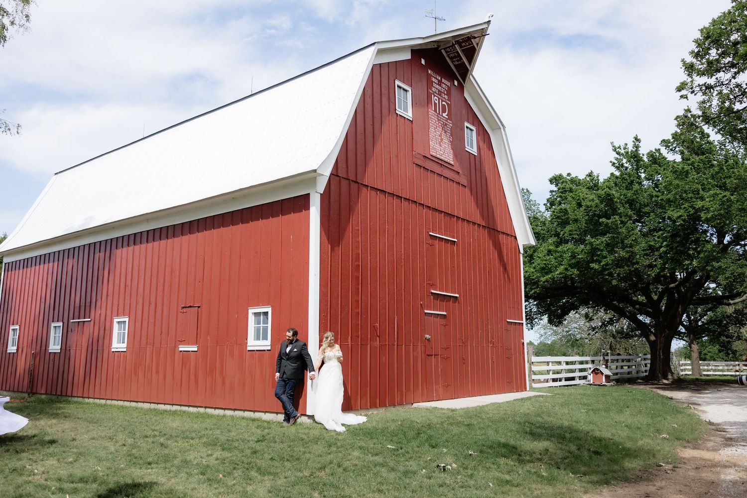 Bride and groom walk hand in hand beside a classic red barn on their rustic outdoor wedding day.