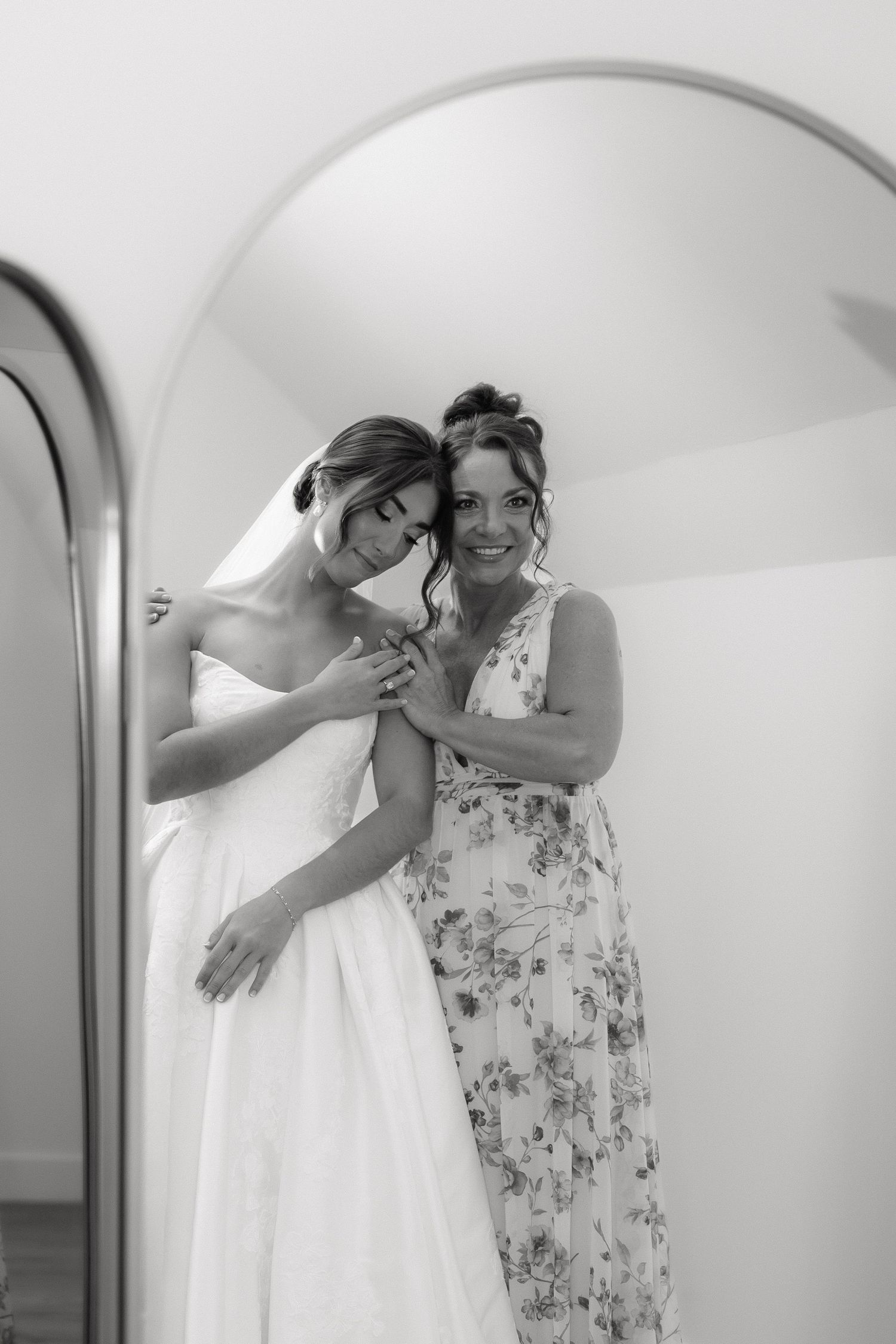 Black and white photo of bride in strapless gown embracing woman in floral dress in mirror.
