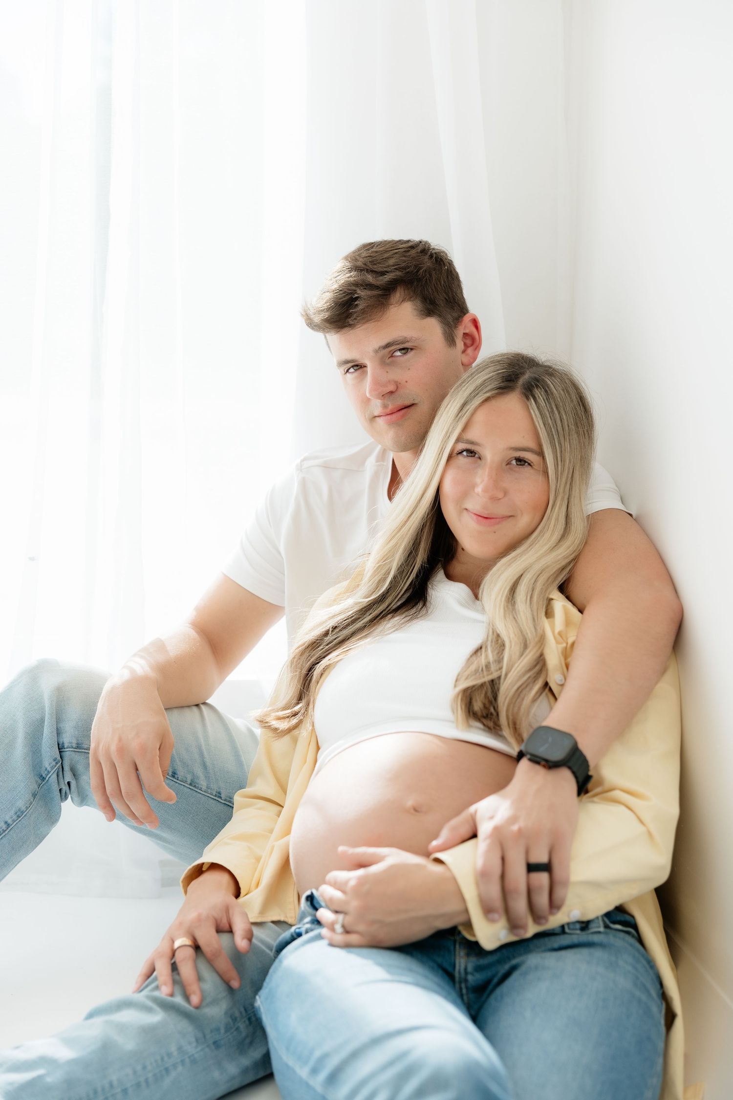 A couple in jeans sits together against a white wall, the man embracing his pregnant partner lovingly.