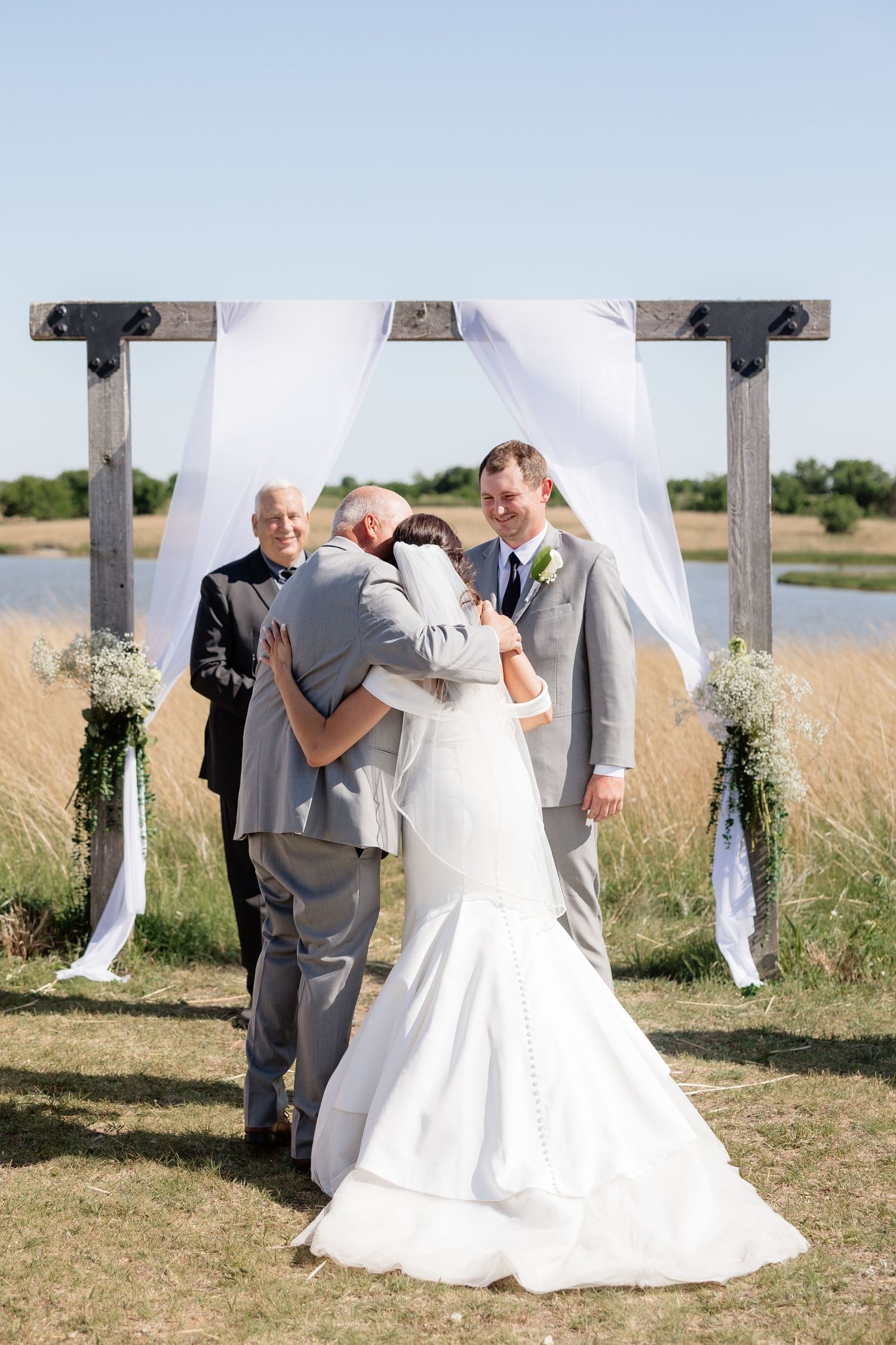 Bride and groom share first hug under rustic wooden arch at outdoor lakeside ceremony.