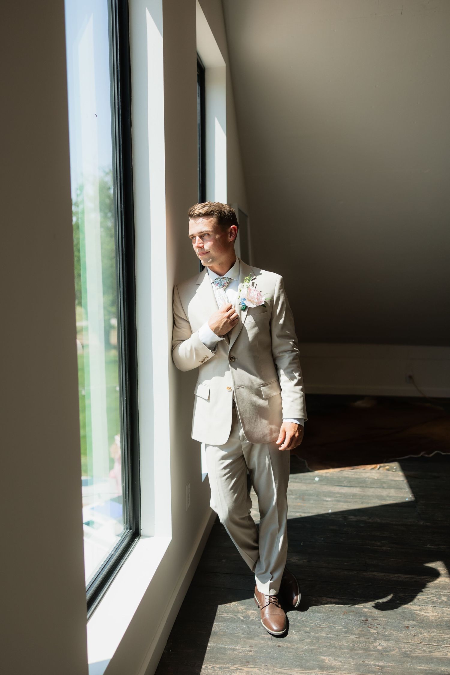 Dapper groom in a tan suit and floral bow tie poses confidently by tall windows in natural light.