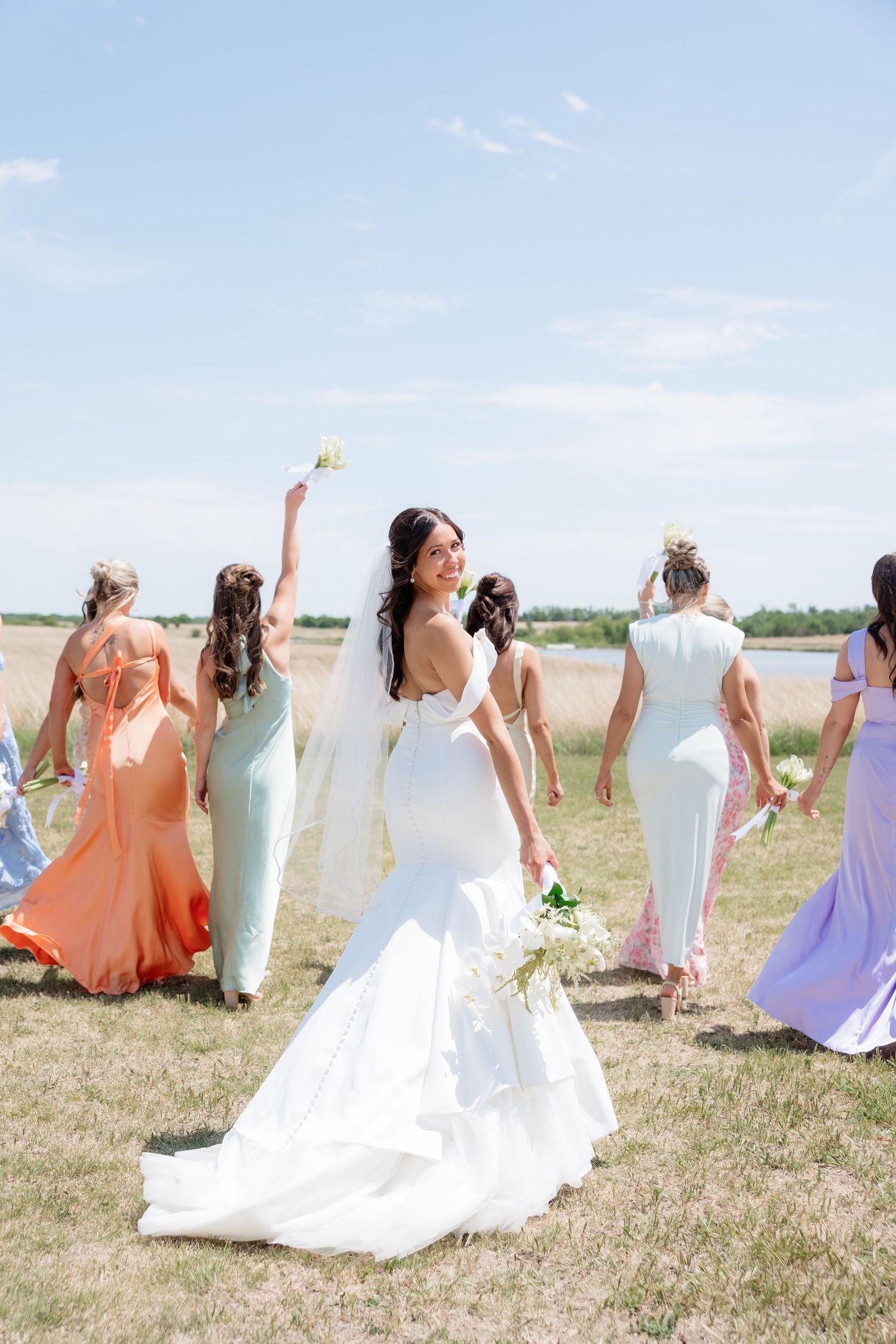 Bride in mermaid gown looks back smiling as bridesmaids walk across sunny field.