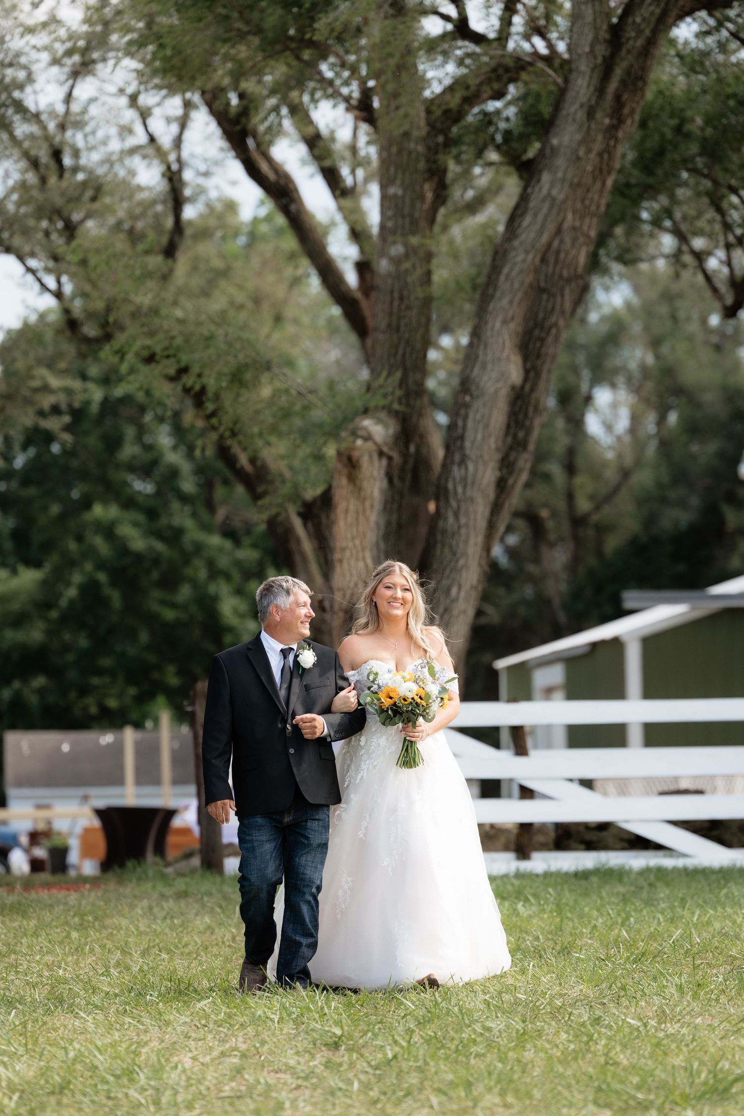 Bride in white gown walks with escort through green outdoor venue before wedding ceremony.