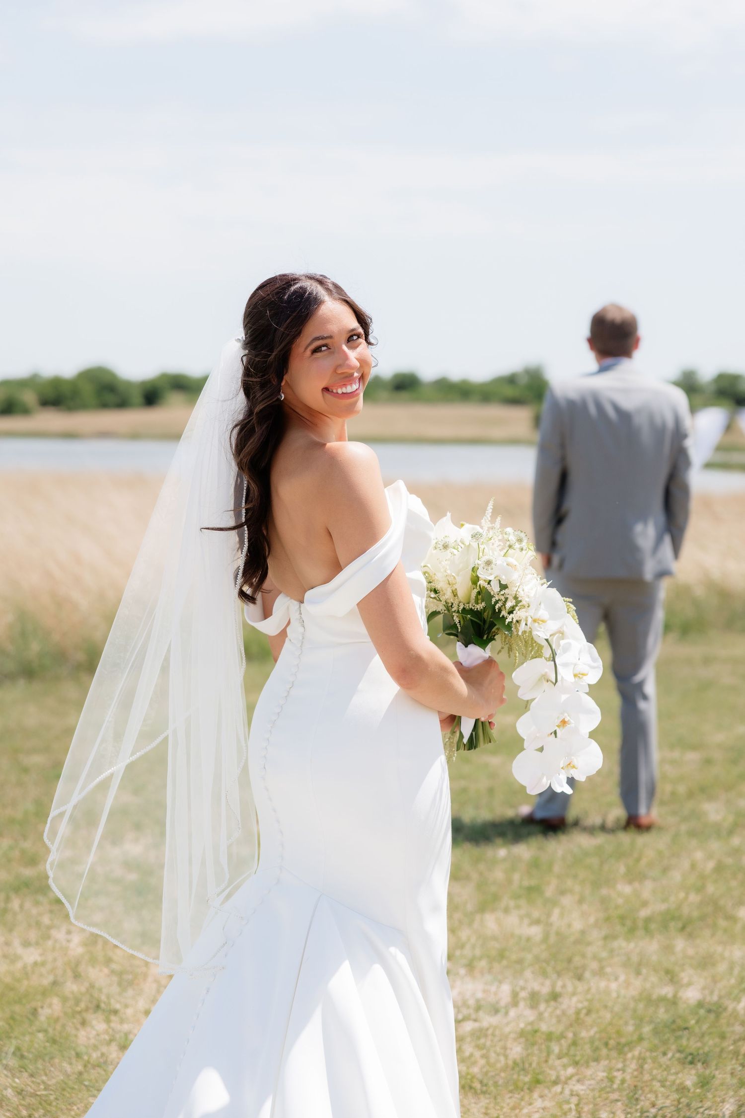 Bride in off-shoulder gown and veil smiles holding white orchid bouquet as groom stands in background.