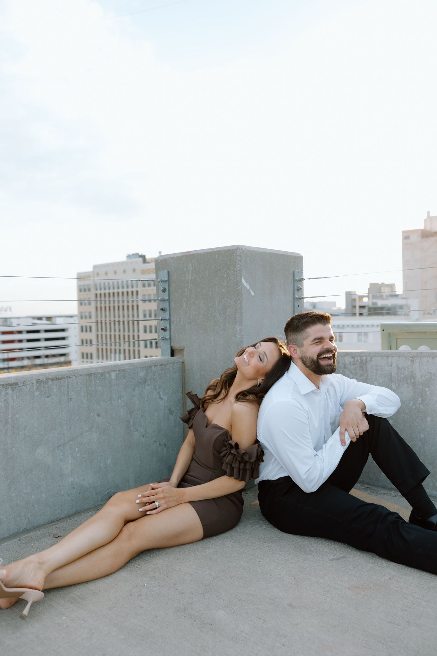 Engaged couple relaxing on urban rooftop, woman in brown mini dress leaning against mans back.