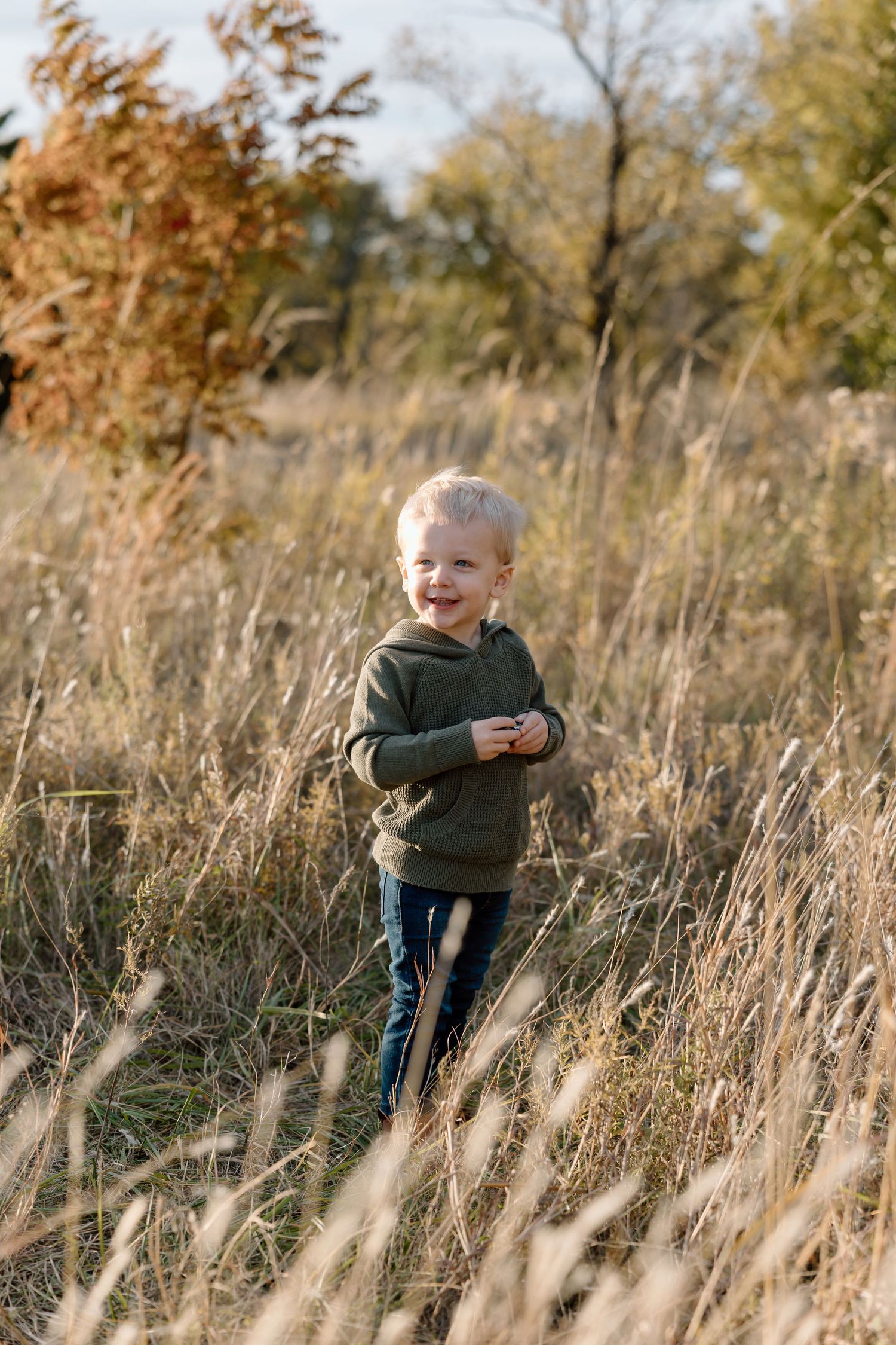 A joyful toddler boy in a green hoodie laughs while playing in tall golden grass during autumn.
