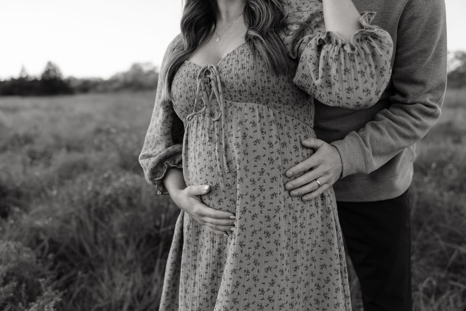 Black and white photo of a couple outdoors, with the man embracing a pregnant woman from behind in a floral dress.
