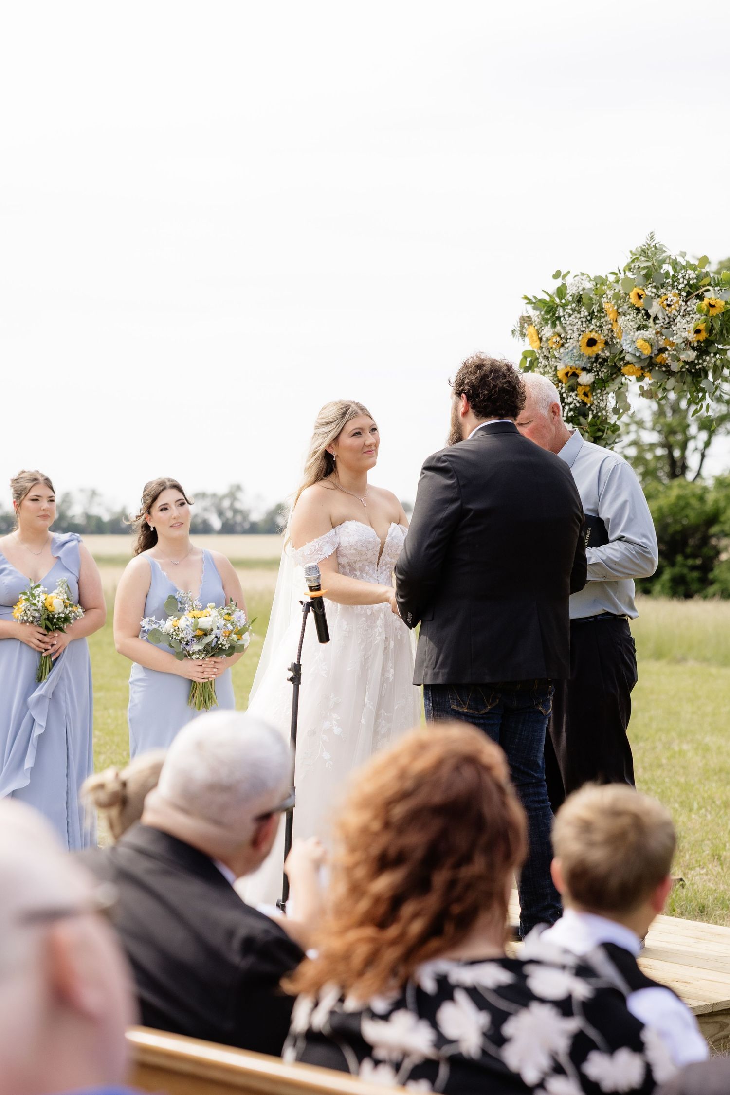 Blonde bride in strapless gown stands at outdoor altar with bridesmaids in blue dresses.