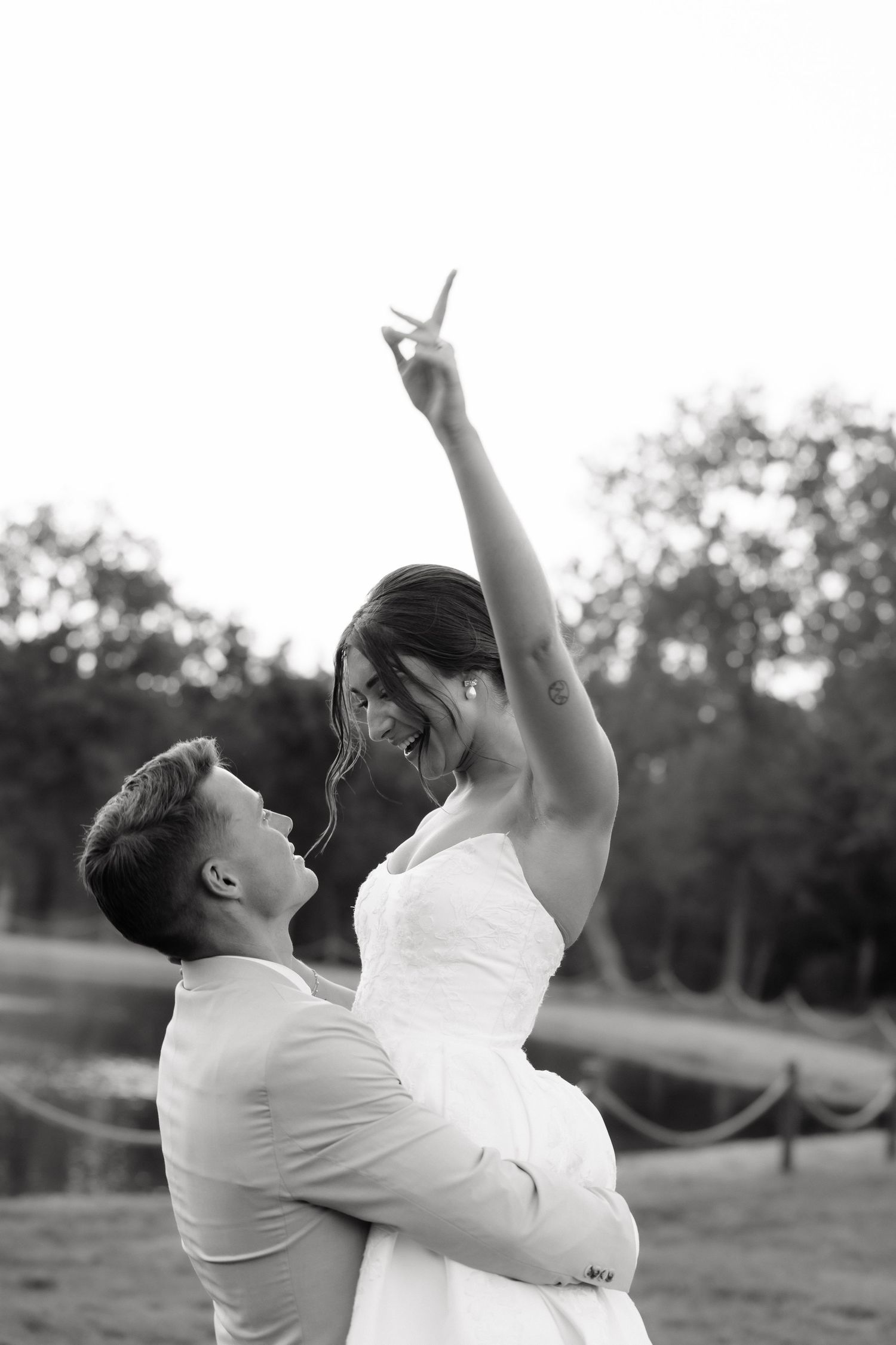 Black and white photo of groom lifting joyful bride outdoors, bride pointing upward near a scenic waterfront.