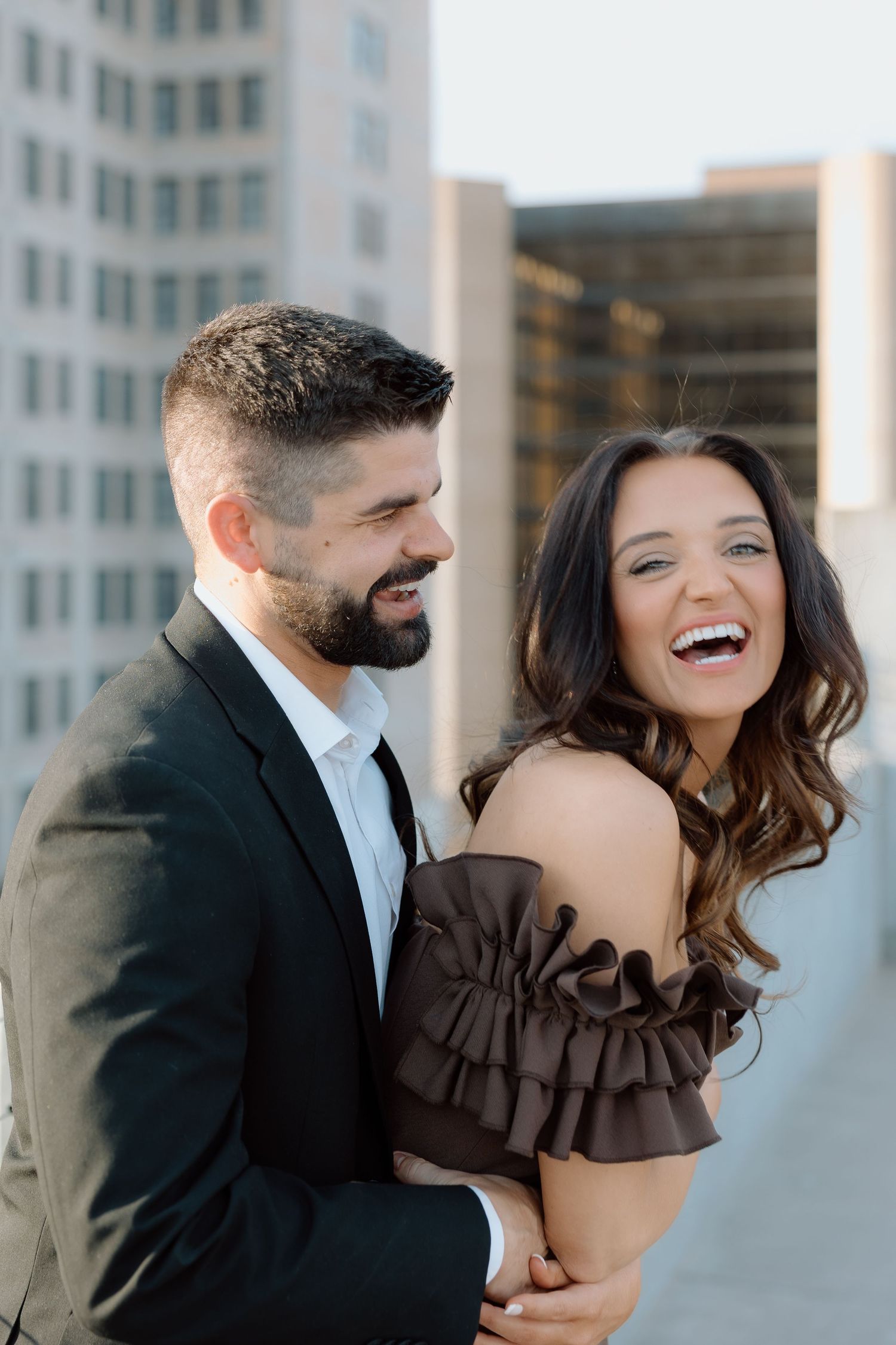 Laughing couple embracing on rooftop with city skyline, woman in brown ruffled off-shoulder dress.
