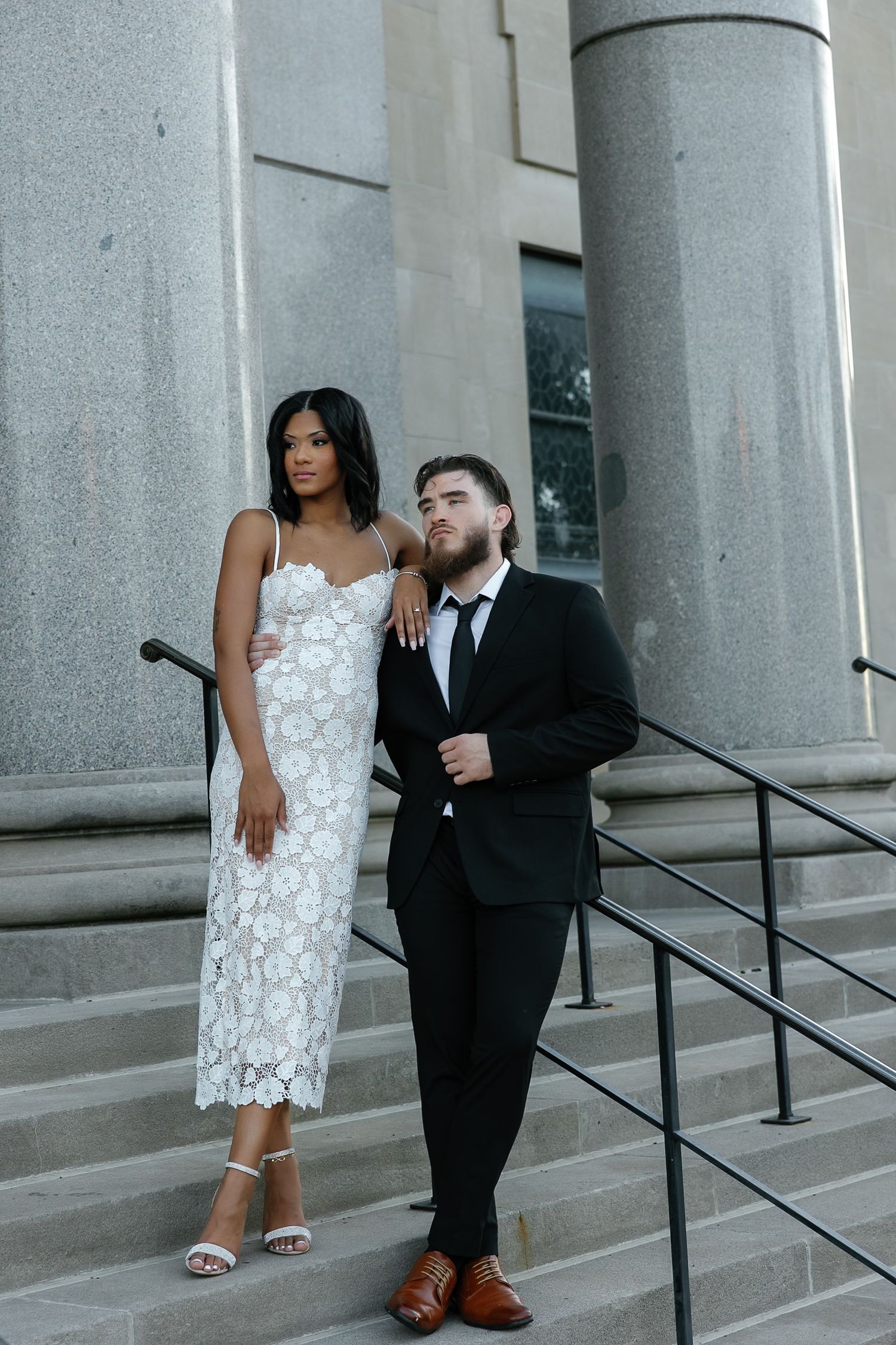 Sophisticated couple posing on grand stone steps, woman in white lace dress and man in black tuxedo.