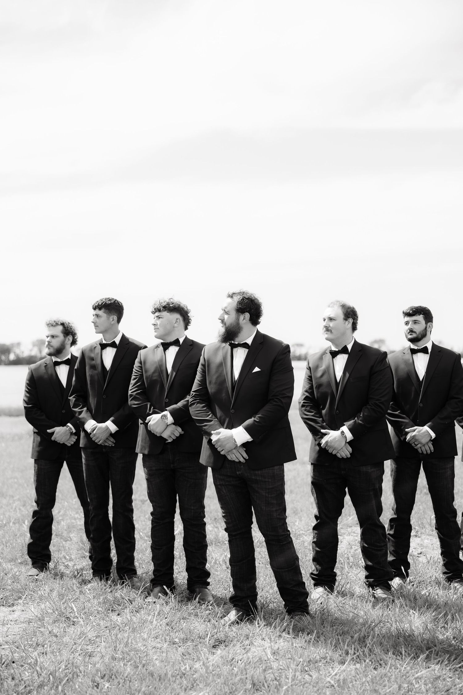 Groom and groomsmen in black tuxedos and bow ties stand in a line in a field in black and white photo.