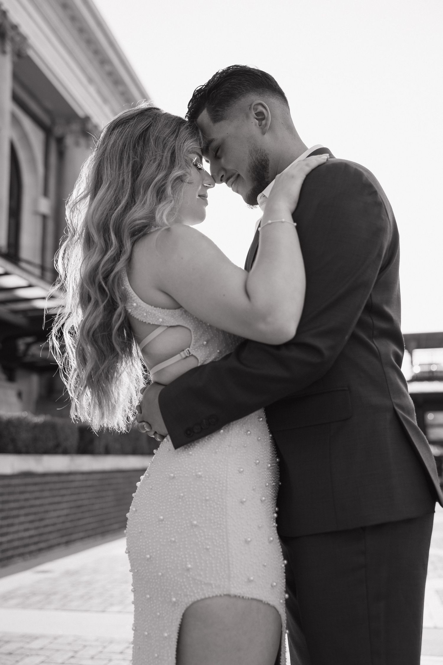 Black and white photo of couple in formal attire embracing forehead to forehead outdoors.