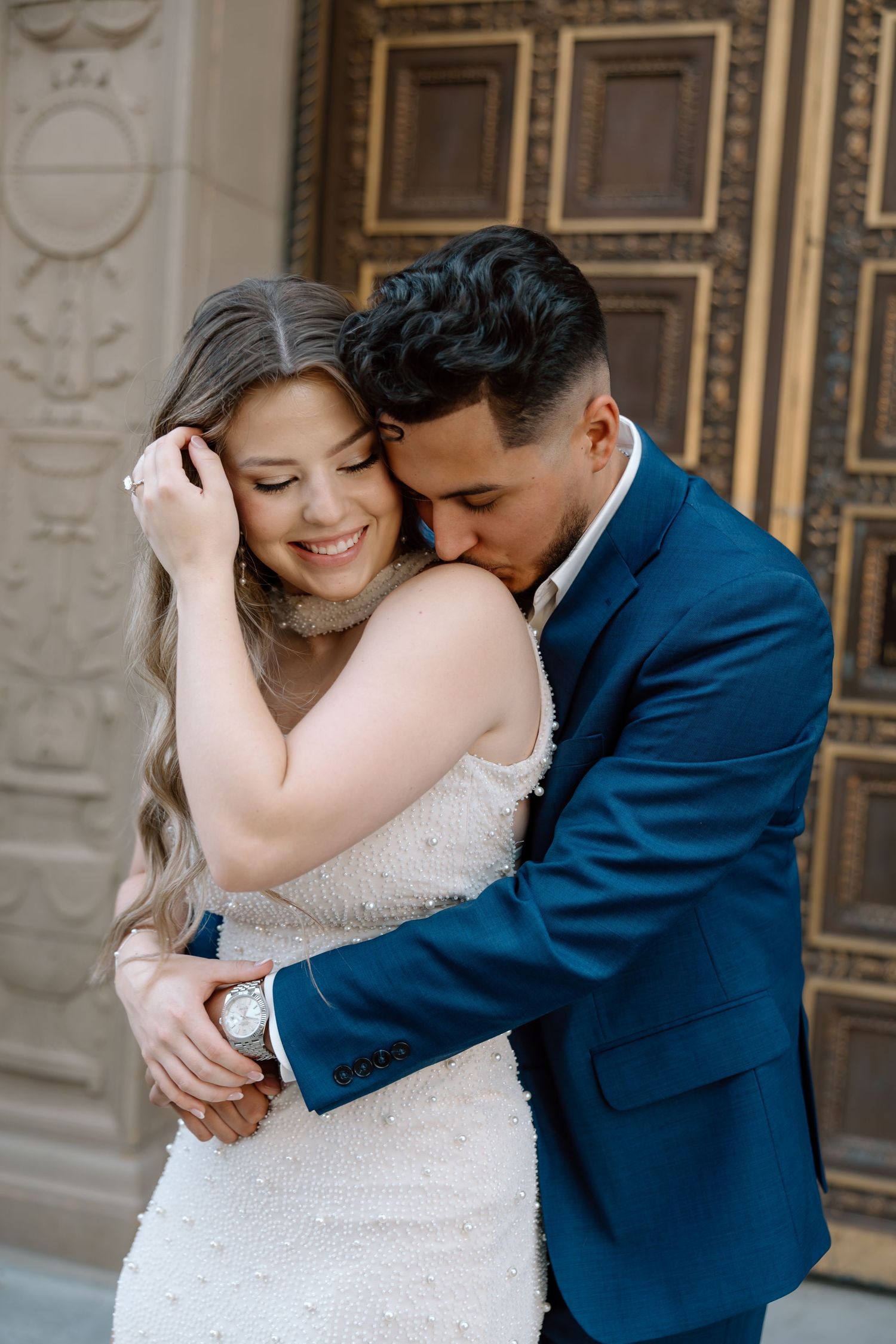 Couple embracing near ornate wooden door, man in blue suit nuzzling woman in white beaded gown.