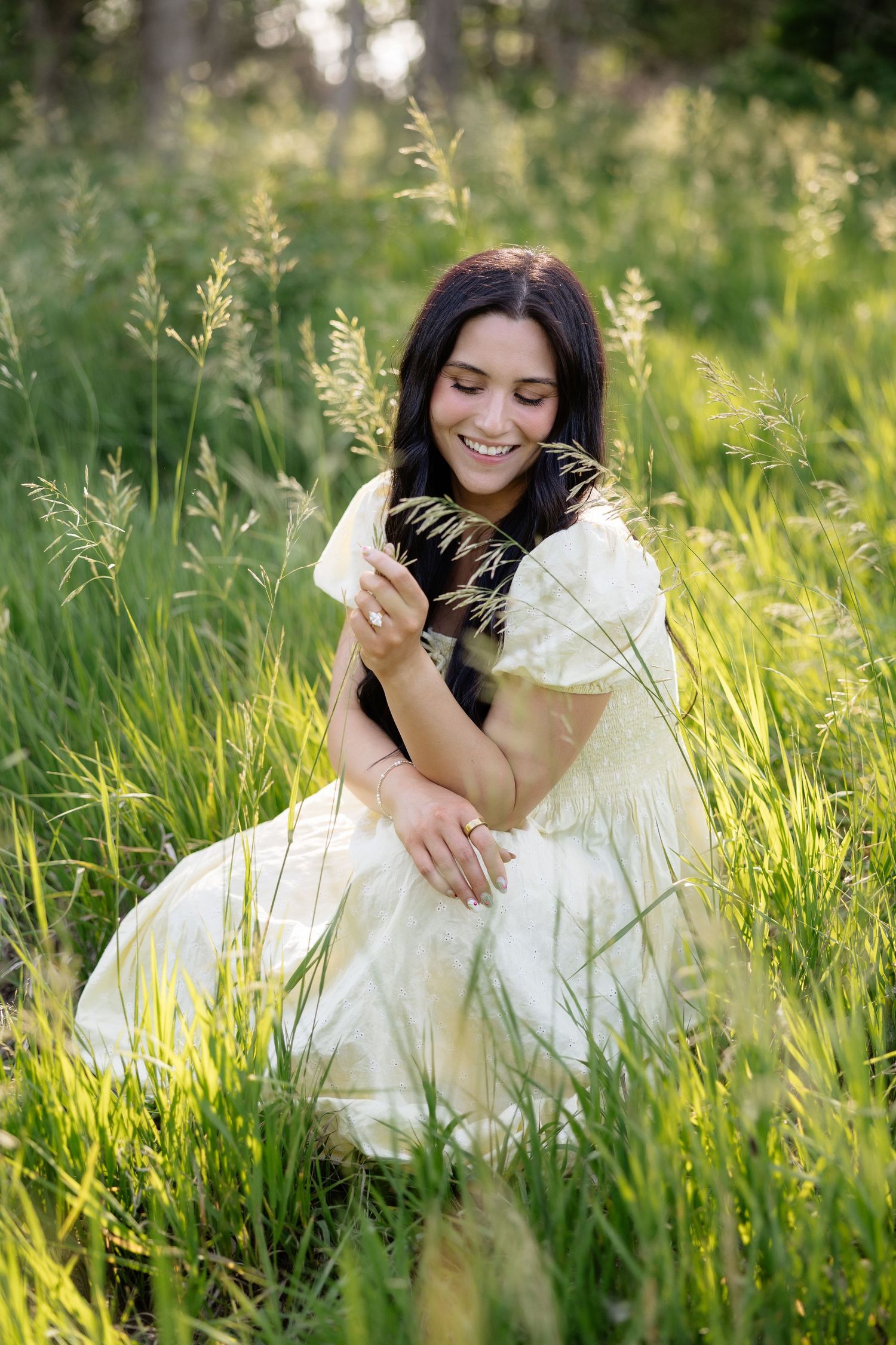 Happy woman in cream dress sitting in tall grass holding a wildflower stem.