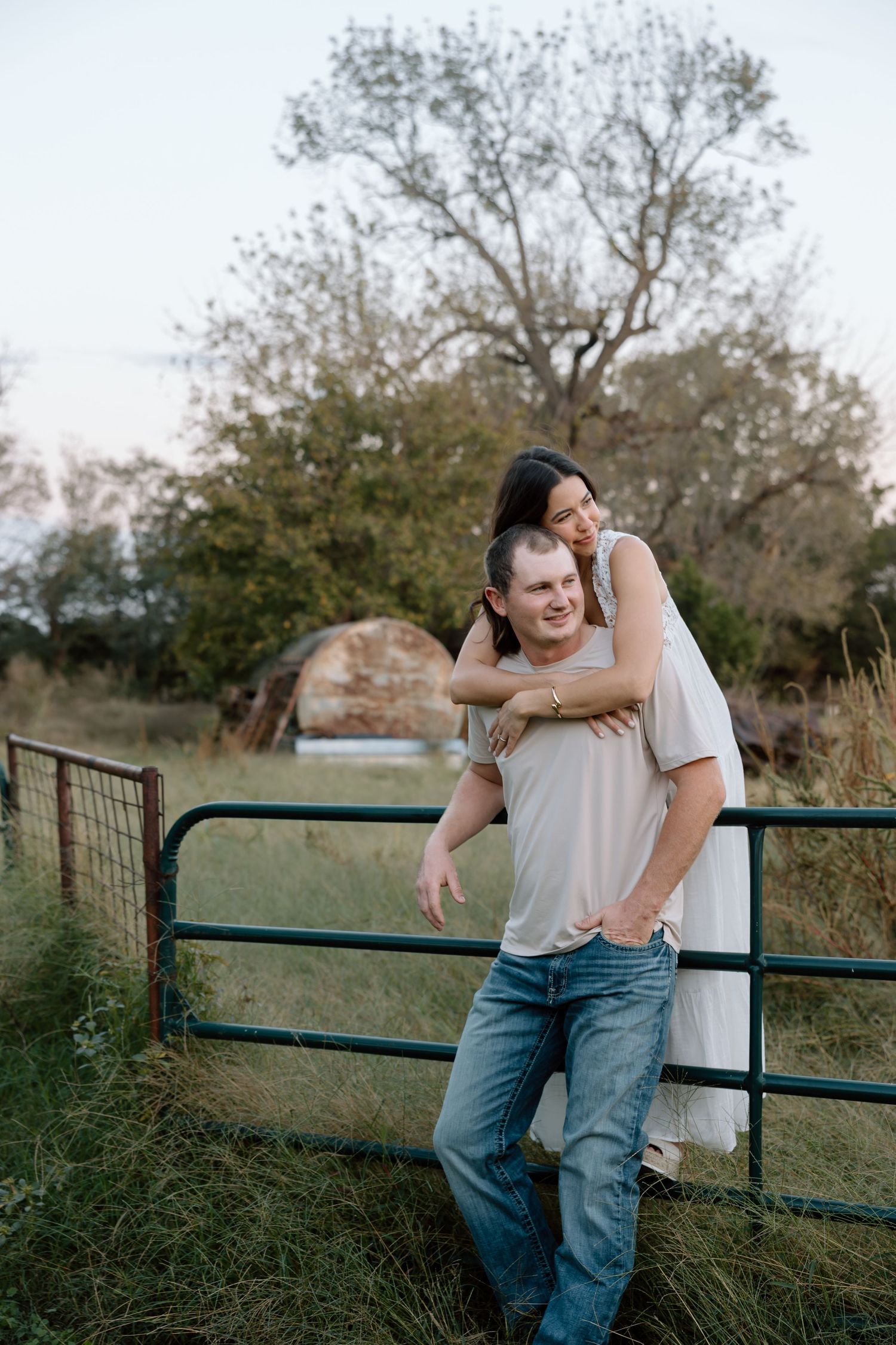 Happy couple posing at a rustic farm gate, woman hugging man from behind in a candid engagement photo.