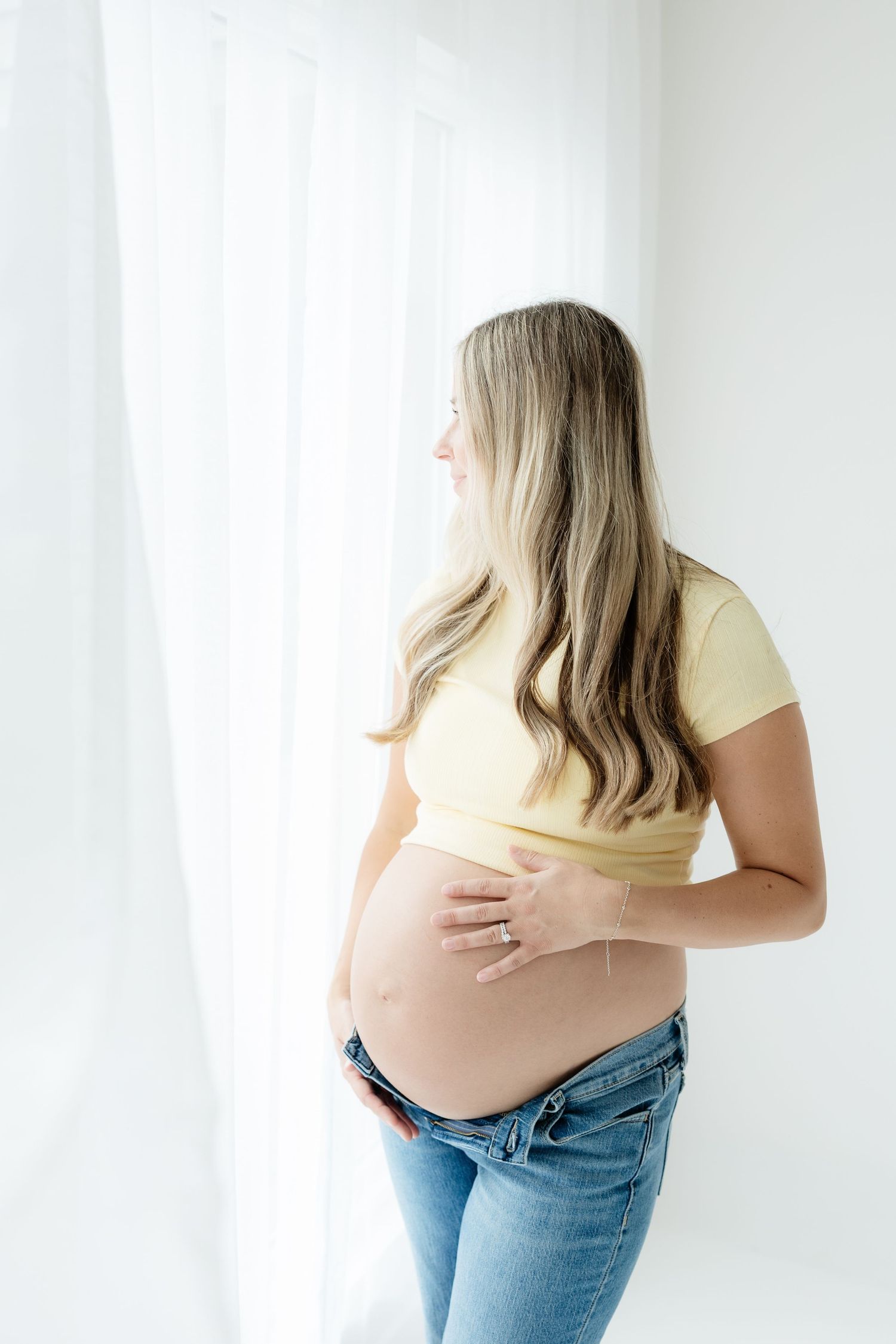 Pregnant woman in yellow crop top and jeans holding baby bump while gazing out a bright window.