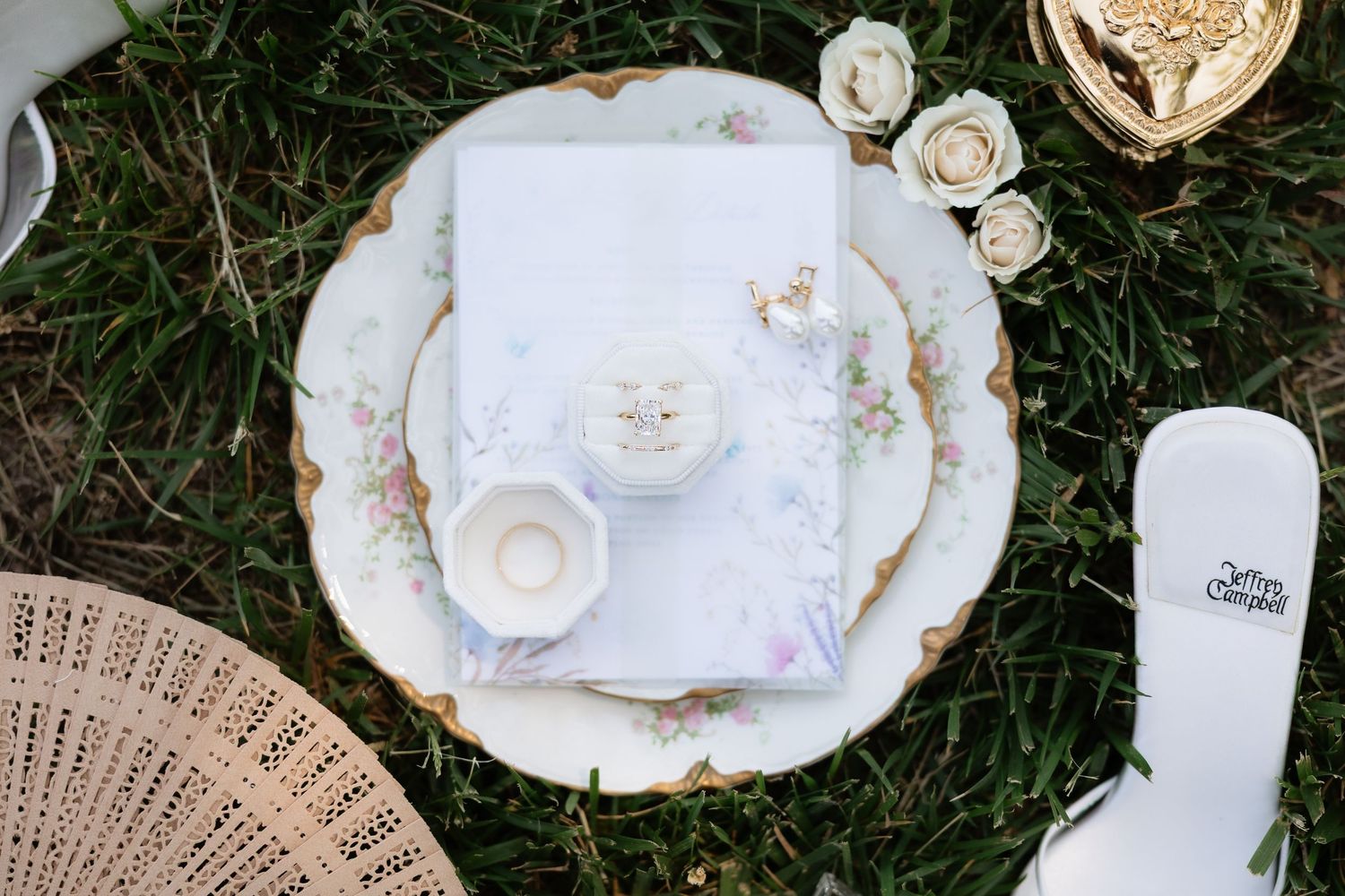 Elegant wedding flat lay with floral plate, vow booklet, candle, and white roses on grass.